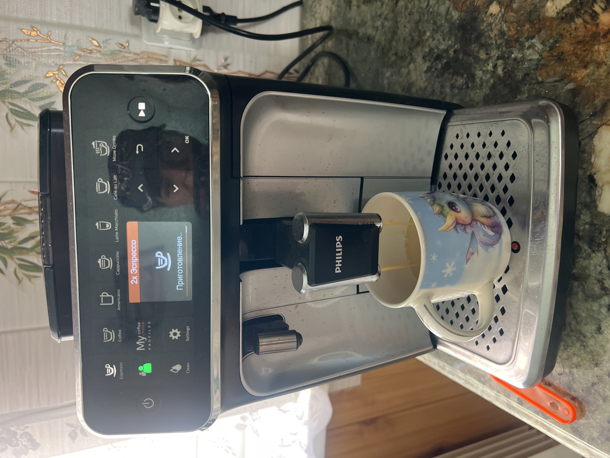 The image shows a Philips coffee machine in the city of Krasnoe-na-Volge, Russia. The machine is actively dispensing coffee into a mug that features a unicorn and snowflakes design. The mug is positioned on the drip tray of the coffee machine. The control panel of the coffee machine displays a selection for "2x Эспрессо" and "Приготовление..", indicating that an espresso is currently being prepared. Various beverage icons and settings are visible on the panel, including Espresso, Coffee, Americano, Cappuccino, Latte Macchiato, Café au Lait, and More Drinks, as well as Clean and Settings. A green indicator light is illuminated next to a silhouette of two people and the text "My coffee PROFILES". The coffee machine is situated on a countertop with a speckled pattern. In the background, a wall with decorative elements and a power outlet with a plug are visible.
