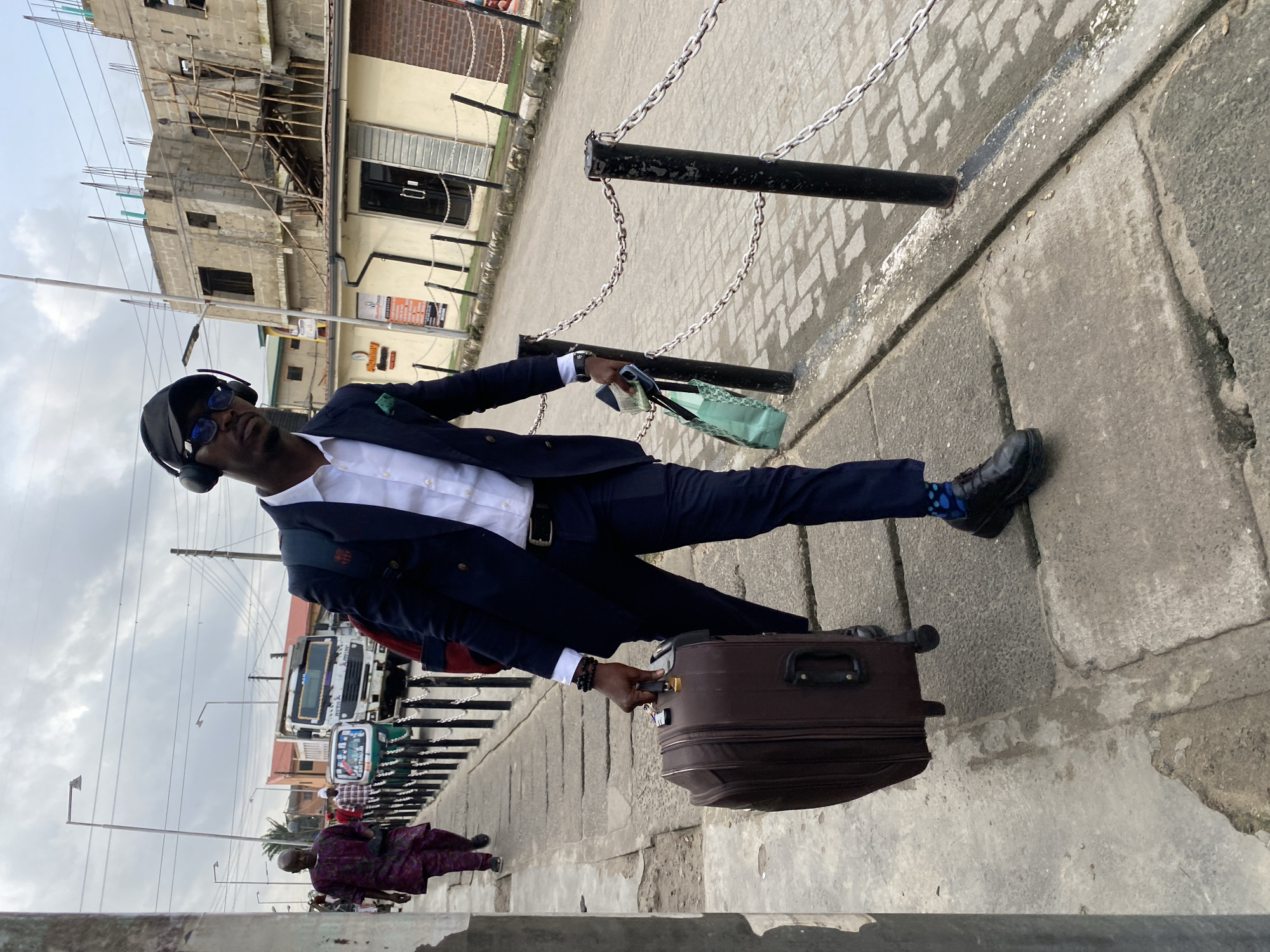 A Black man wearing a dark suit, white shirt, black cap, sunglasses, and headphones is walking on a sidewalk, carrying a dark brown suitcase in his right hand and a light green shopping bag in his left. He is also wearing patterned blue socks and black dress shoes. To his left, another Black man in a purple patterned traditional garment is walking away from the camera. In the background, there is a truck, a three-wheeled vehicle, a building under construction, and other structures. The scene appears to be an outdoor urban environment with a cloudy sky.