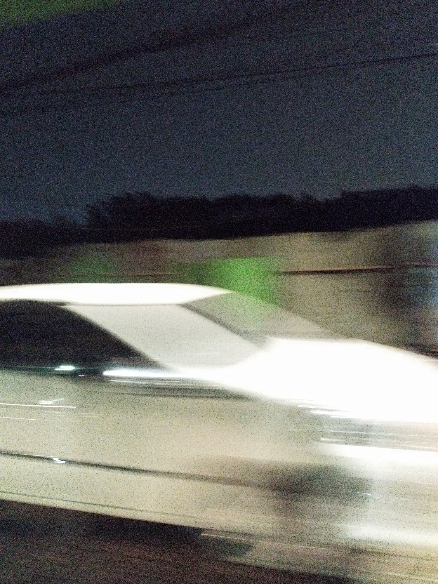 The image depicts a blurred, night-time scene of a car in motion on a road. The car's headlights create streaks of light, indicating a long exposure. The background shows a dark, silhouetted tree line against a dark sky, with a faint green glow in the mid-ground. The location is identified as a road in Surabaya, Indonesia.