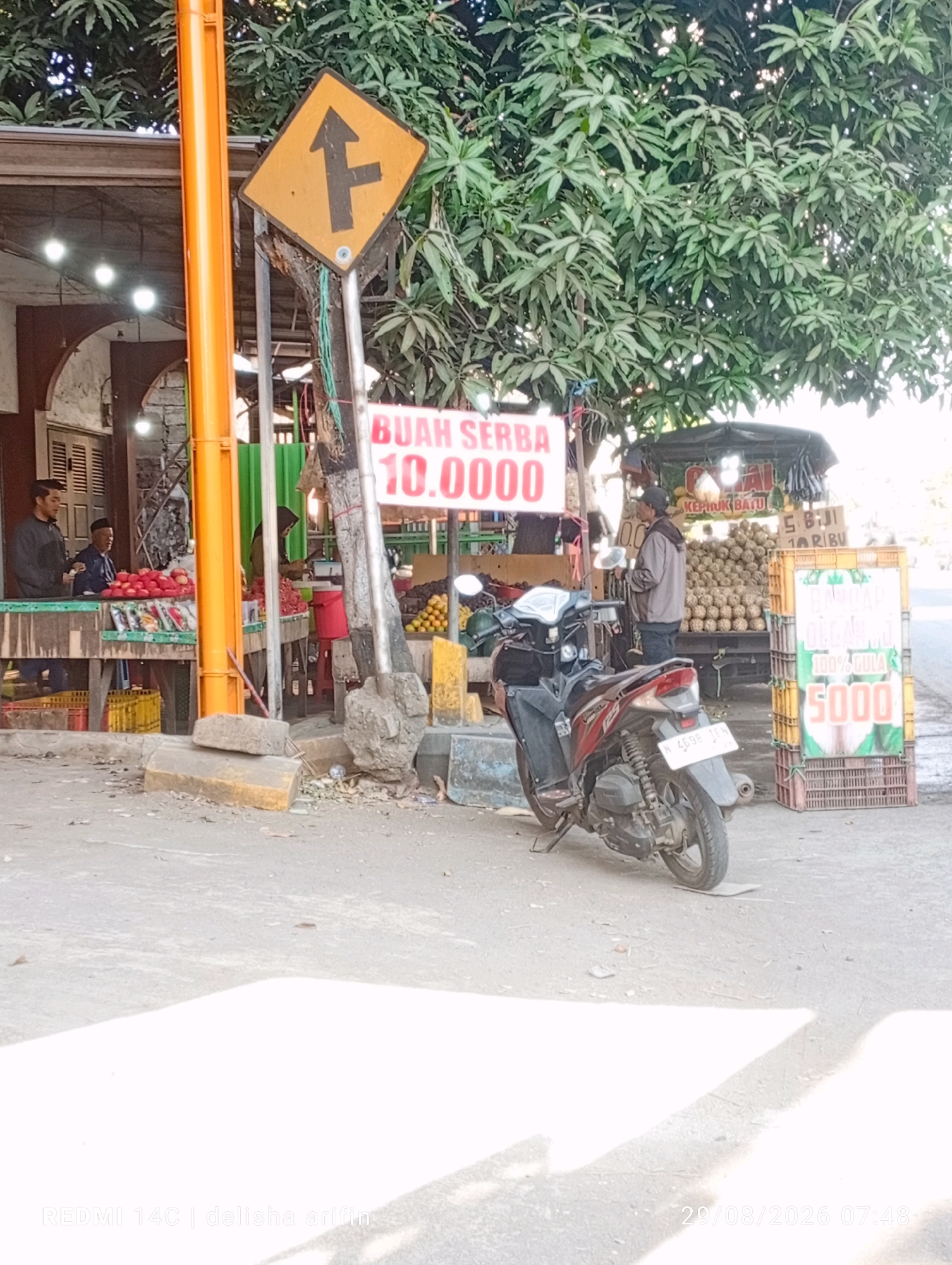 The image depicts a street-level view of a small market or roadside stall in Wonorejo, Indonesia, on a sunny day. In the foreground, a parked red and black scooter faces towards the right. The background features a market stall with various fruits displayed, including what appears to be custard apples on a truck and possibly tomatoes on a table. Several people are visible, including a man in a grey jacket interacting with a stall, and two men standing at a stall selling red produce. A traffic sign indicating a lane split is present, positioned near a bright orange pole. The environment is a paved area with scattered debris, suggesting an outdoor, somewhat informal commercial setting. The lighting indicates it is daytime, with strong shadows cast on the ground.