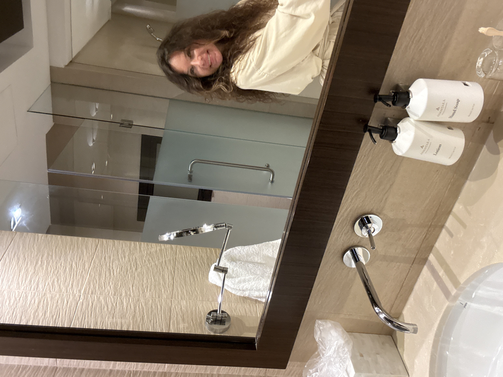 The image is a selfie taken in a bathroom, likely in a hotel in Dubai, United Arab Emirates. A woman with long, curly brown hair is reflected in a large mirror, wearing a light-colored robe. She is smiling and looking towards the camera. To her left in the reflection, a glass shower enclosure with a silver handle is visible, along with a wall-mounted towel rack holding a white towel. In the foreground of the actual image, a modern bathroom sink and faucet are visible, along with two white pump bottles labeled "Lotion" and "Hand Soap." The walls of the bathroom are tiled with a textured, light beige material.