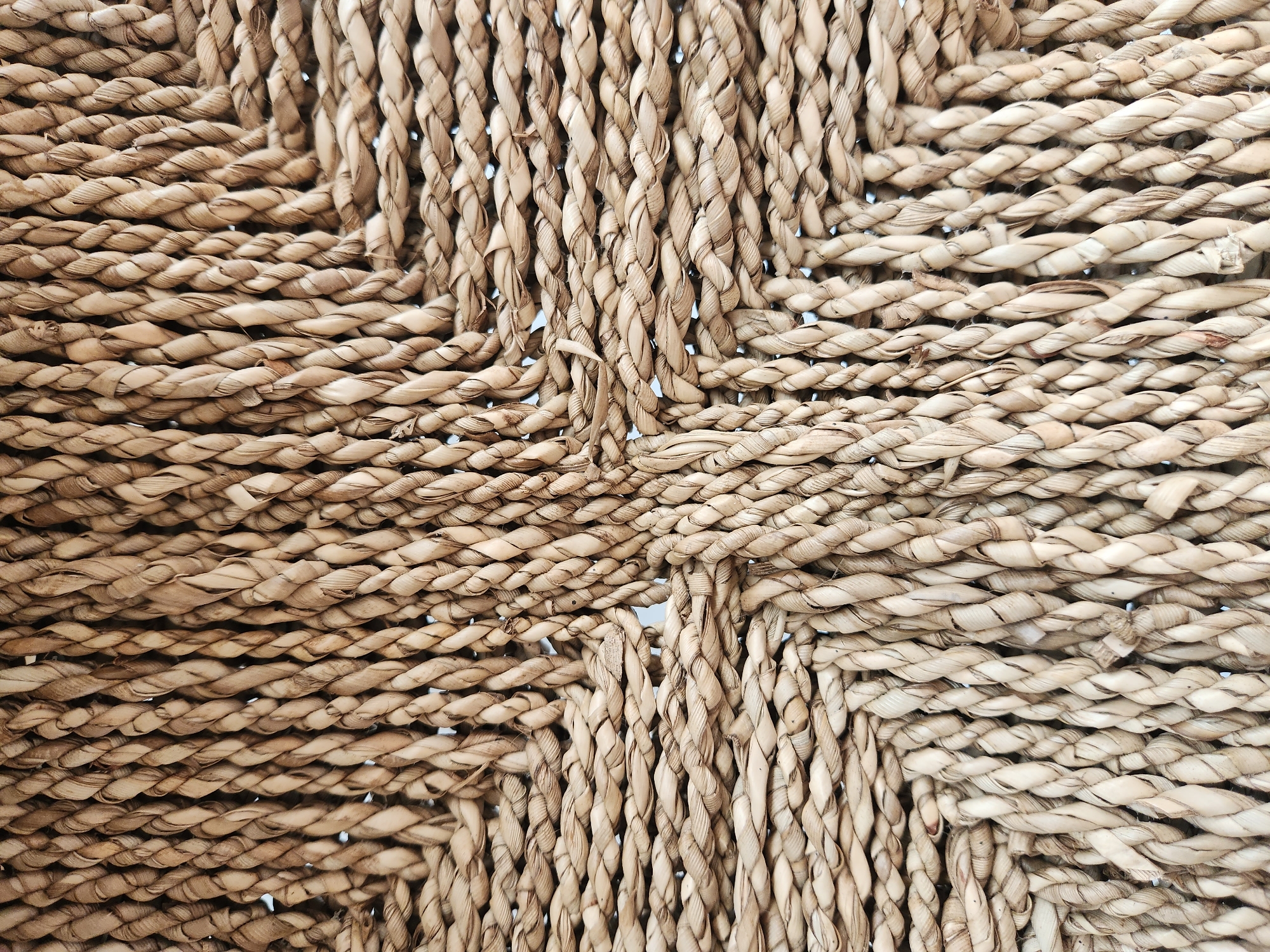 This is a close-up, full-frame shot of a woven material, likely from a basket or furniture. The texture is the dominant subject, with thick, braided strands of natural fiber, possibly straw or rattan, creating a crisscross pattern. The fibers are a light, earthy brown with variations in tone, suggesting they are dried and natural. The weave is dense and appears to be a basket weave, with vertical and horizontal strands interlocked. The lighting is even, highlighting the texture and subtle shadows within the weave, suggesting it's well-lit, possibly in daylight. No people, animals, or specific setting are visible; the focus is solely on the intricate texture of the woven material. The overall impression is one of natural material and handcrafted construction. There is no visible text. The location context is Maitland, United States of America.