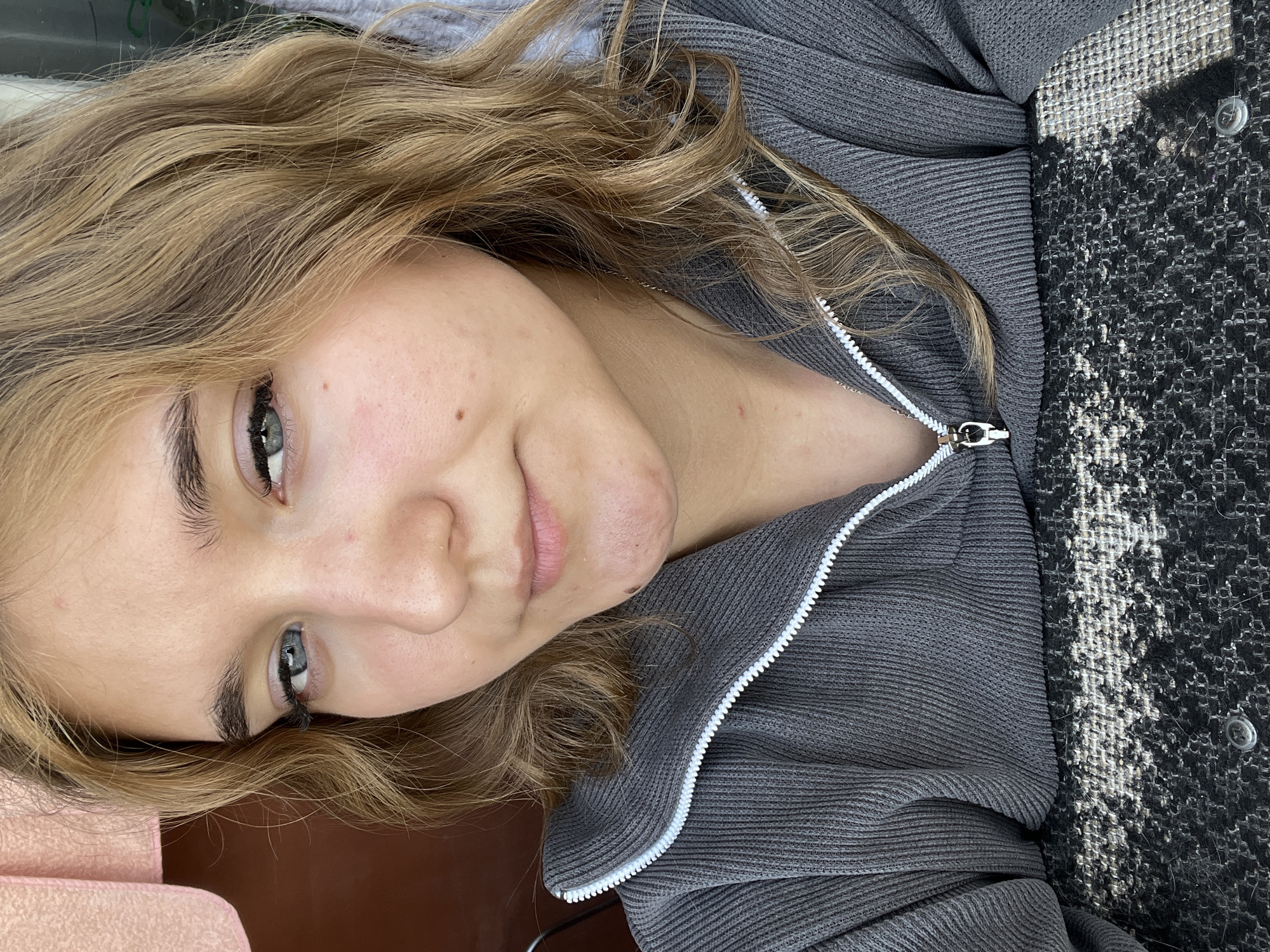 A young woman with blonde, wavy hair and blue eyes is pictured in a close-up, head-and-shoulders shot. She is wearing a grey, ribbed zip-up jacket. Her skin has some redness and a few blemishes. The background is blurred, with hints of pink fabric on the left and a dark brown surface behind her. The overall lighting suggests an outdoor setting, possibly in Chaikovsky, Russia, given the prompt.