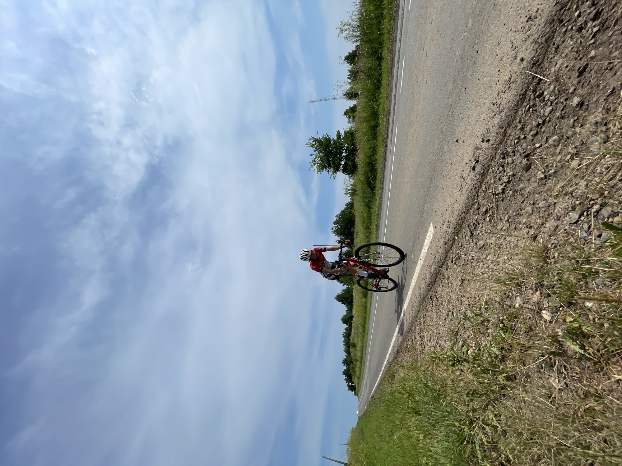 An individual wearing a red and white cycling suit and a white helmet is riding a red road bicycle on a paved asphalt road. The cyclist is positioned in the middle of the right lane, facing away from the camera and towards the left of the frame. The road extends into the distance and curves slightly to the left. On the left side of the road, there is a grassy embankment with some dry vegetation. The right side of the road is bordered by a grassy area with scattered trees and wildflowers. In the background, there is a line of trees, and further in the distance, a tall, slender radio tower with a red top is visible. The sky is blue with scattered white clouds. The scene appears to be set in a rural or suburban area, possibly the village of Perevoznoe, Russia, given the presence of a radio tower common in such locations. The lighting suggests it is daytime, with the sun casting shadows.
