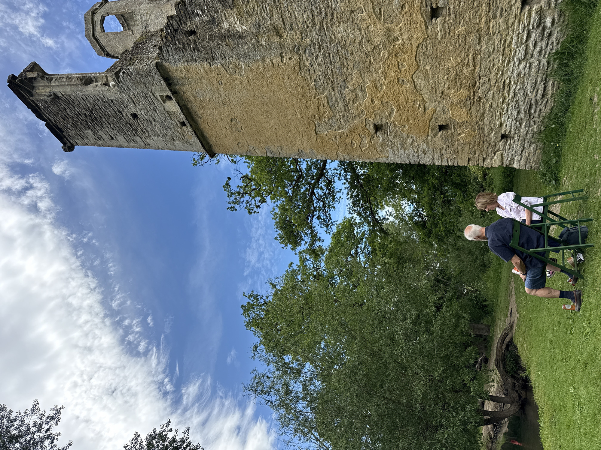 The image shows a tall stone structure, likely a tower or part of a castle ruin, in West Oxfordshire, United Kingdom. The structure is made of rough-hewn stones and shows signs of age and weathering. It rises high against a blue sky with scattered clouds. At the base of the structure, in a grassy area, two people are sitting on green chairs. One person is older with white hair, wearing a dark shirt and shorts. The other person, possibly a woman, is wearing a white top. They appear to be relaxing or enjoying the view of the stone structure amidst the surrounding greenery, including various trees and foliage. There is a stream with an exposed tree root system winding nearby.