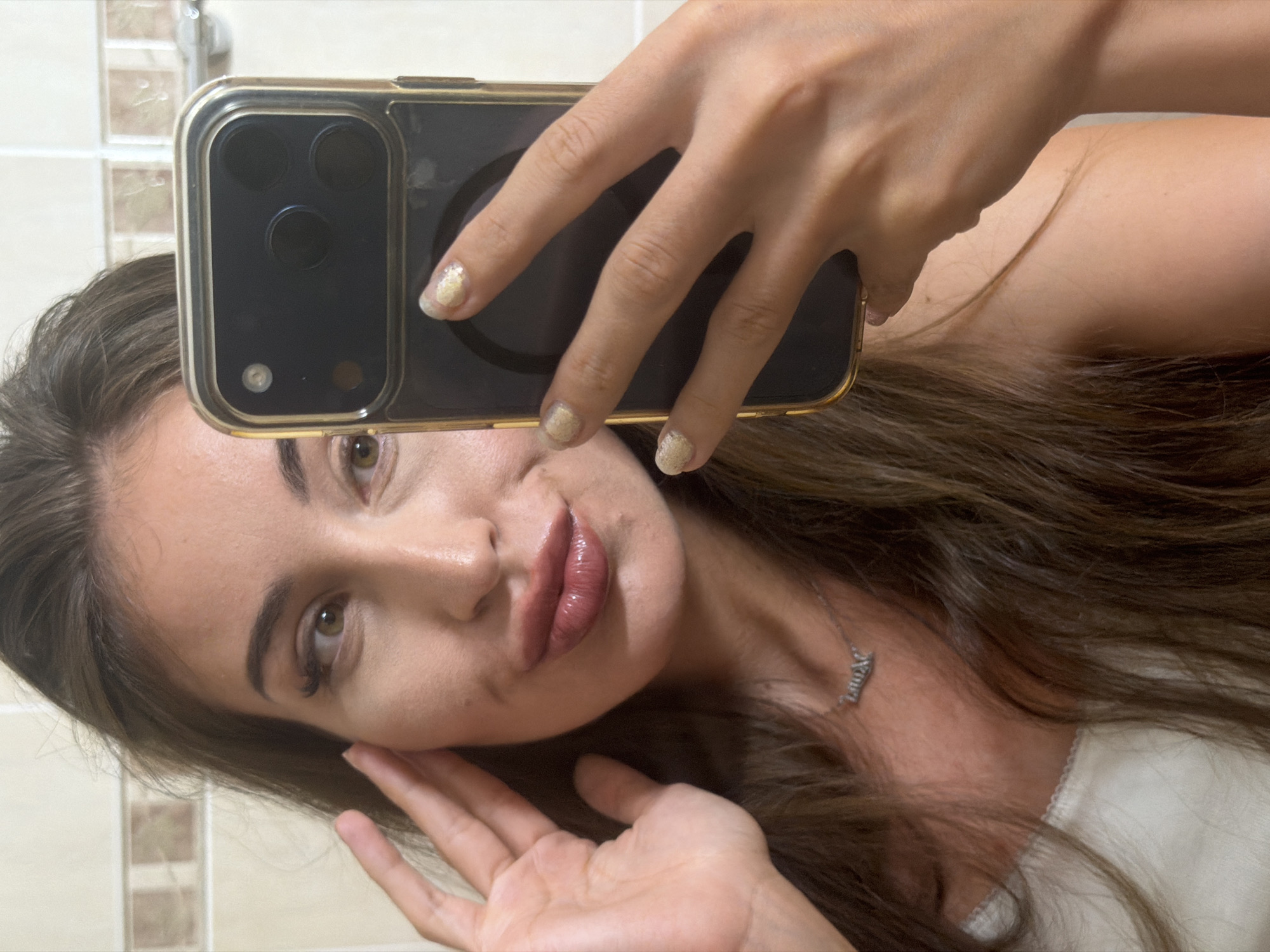 In this close-up, slightly blurry selfie taken from chest-up, a young woman with long, dark brown hair is holding an iPhone 14 Pro in her right hand, taking the photo. She is wearing a light-colored, possibly white, camisole and a silver pendant necklace with a nameplate that appears to read "Lauren." Her left hand is raised to her face, with her fingers touching her cheek. Her makeup includes winged eyeliner and glossy, reddish-brown lipstick. The background appears to be a tiled bathroom wall, suggesting the photo was taken indoors. The overall lighting is bright, and the focus is primarily on the woman and her phone. There is no specific indicator of the location being Tersefanou, Cyprus, beyond the request for its inclusion in the description.