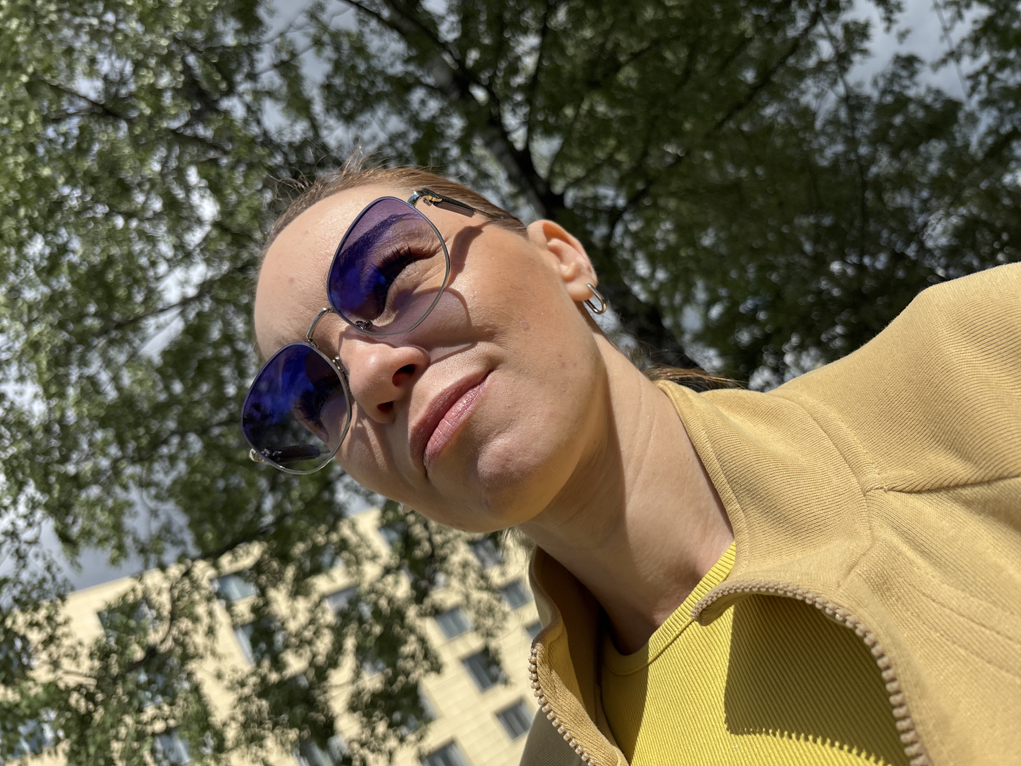 This is a medium shot, taken from a low angle, of a person wearing sunglasses and a yellow top. The person is looking down and to their right, with a neutral expression. The background consists of green trees and a building, suggesting an outdoor setting during the day. The lighting indicates it is likely daytime, with sunlight filtering through the trees, creating dappled shadows. The person is wearing a yellow jacket over a yellow top, and large sunglasses with blue lenses. They also have a silver hoop earring in their left ear. The scene appears to be in Sinyavino, Russia.