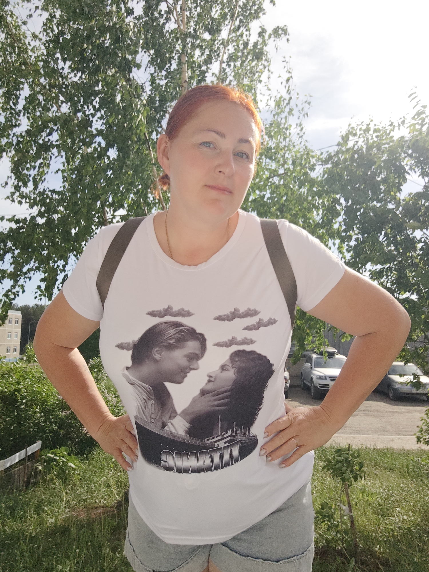 A woman with reddish hair stands outside in Markovskii, Russia, posing with her hands on her hips. She is wearing a white t-shirt featuring an image of Jack and Rose from the movie "Titanic," along with faded denim shorts. A backpack strap is visible on her shoulder. Behind her are trees, grass, a street, parked cars, and a building. The sky is bright, suggesting it is daytime.