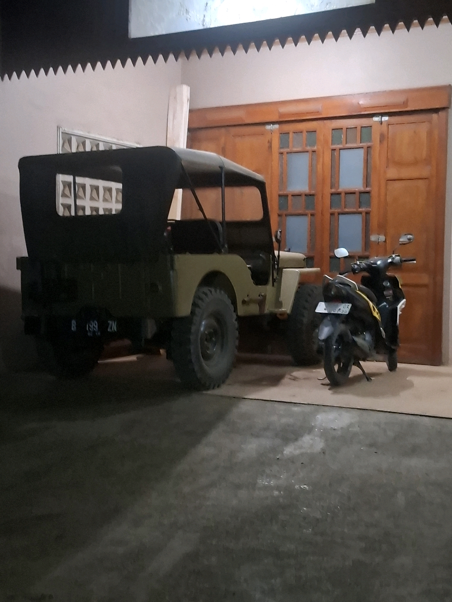 The image depicts a street scene at night in Ciheulang Tonggoh, Indonesia. In the foreground, a vintage olive green jeep with a black soft top is parked. The jeep has a license plate that reads "B 199 ZN". To the right of the jeep, a black motorcycle with yellow accents is parked. The vehicles are situated on a concrete surface, possibly a driveway or a street, in front of a building with a large wooden sliding door. The building has decorative white trim along the top edge. The lighting suggests it is nighttime, with artificial light illuminating the scene.