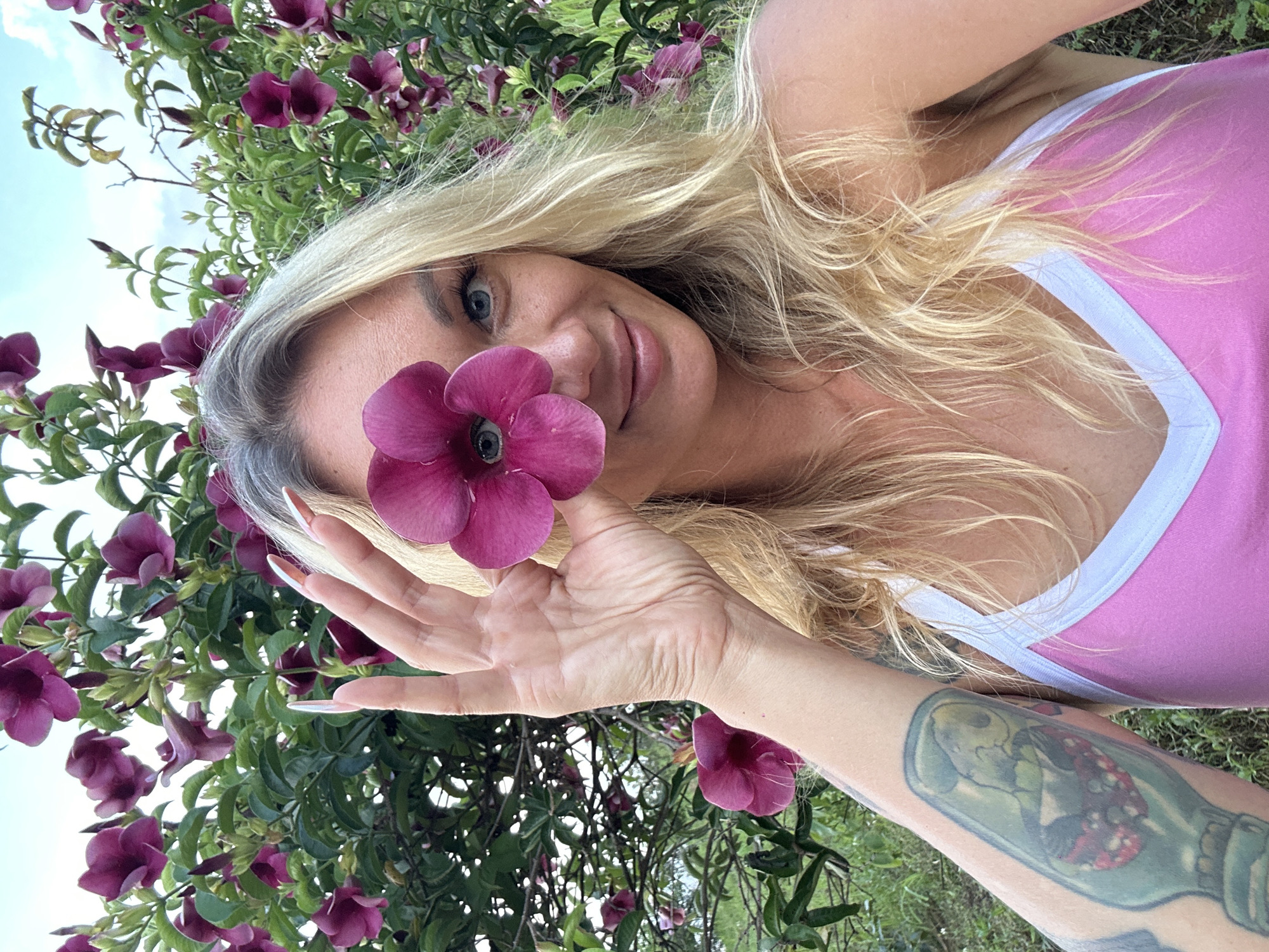 A woman is captured in a close-up, horizontal shot, holding a deep pink flower to her face, partially obscuring it. She has blonde hair that cascades around her face and shoulders. Her eyes are a striking blue, and she is smiling gently. She is wearing a pink tank top with white trim. Her right hand is visible, with long, manicured nails, positioned near the flower. A tattoo is visible on her left forearm, depicting what appears to be a figure inside a bottle surrounded by floral elements. The background is filled with lush green foliage and more deep pink flowers, suggesting an outdoor setting, possibly a garden. The lighting indicates it is daytime. The overall impression is one of natural beauty and serene enjoyment.
