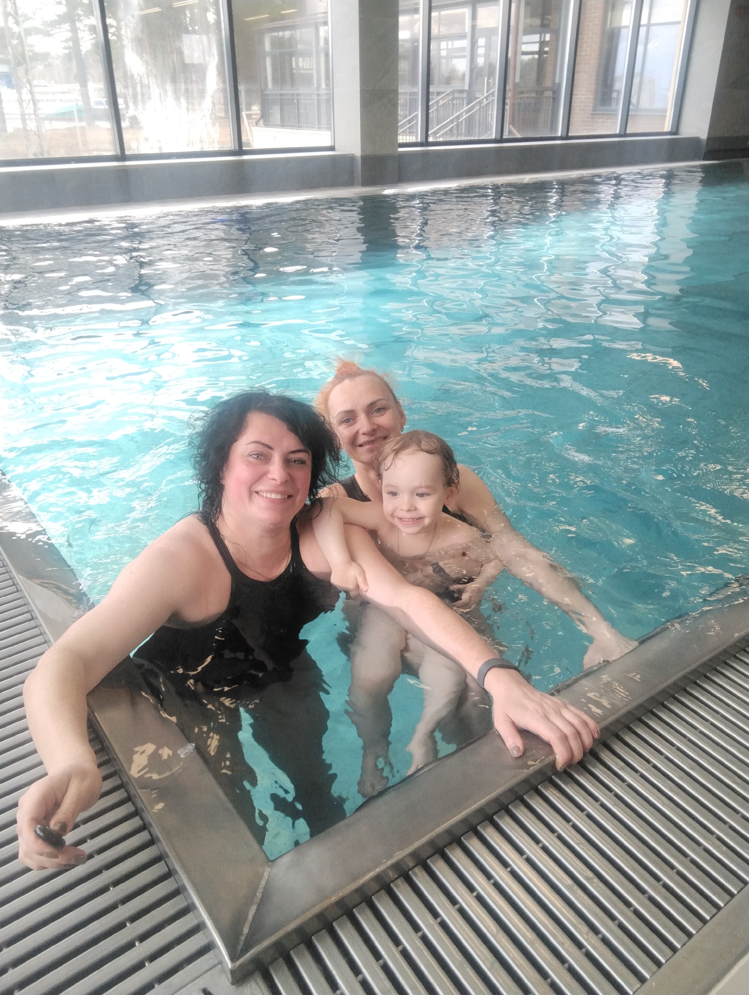 This is a brightly lit, indoor portrait featuring three individuals – two adult women and one young child – enjoying themselves in a swimming pool in Chaikovsky, Russia. The pool water is clear and light blue, with gentle ripples on its surface.
On the left, an adult woman with dark, curly hair and a black swimsuit smiles warmly at the camera. She is leaning against the pool's edge, gripping its metallic border with both hands. Her left hand shows dark nail polish.
Positioned behind her and slightly to the right, another adult woman with reddish-blonde hair also smiles cheerfully at the camera. She is holding and supporting a young child, who appears to be a toddler or preschooler, with light brown hair and a wide, joyful smile. The child looks happily at the camera while being held in the water. The second adult woman wears a dark wristband on her left wrist.
In the background, large floor-to-ceiling windows reveal an outdoor scene featuring what appears to be a modern building with multiple windows and some trees, indicating a daytime setting with ample natural light. The pool's perimeter includes a gray, slatted drainage system.
The overall atmosphere of the image is joyful and relaxed, with all subjects displaying happy and contented expressions, suggesting a pleasant time spent together in a leisure or recreational pool environment. There is no visible text in the image.
