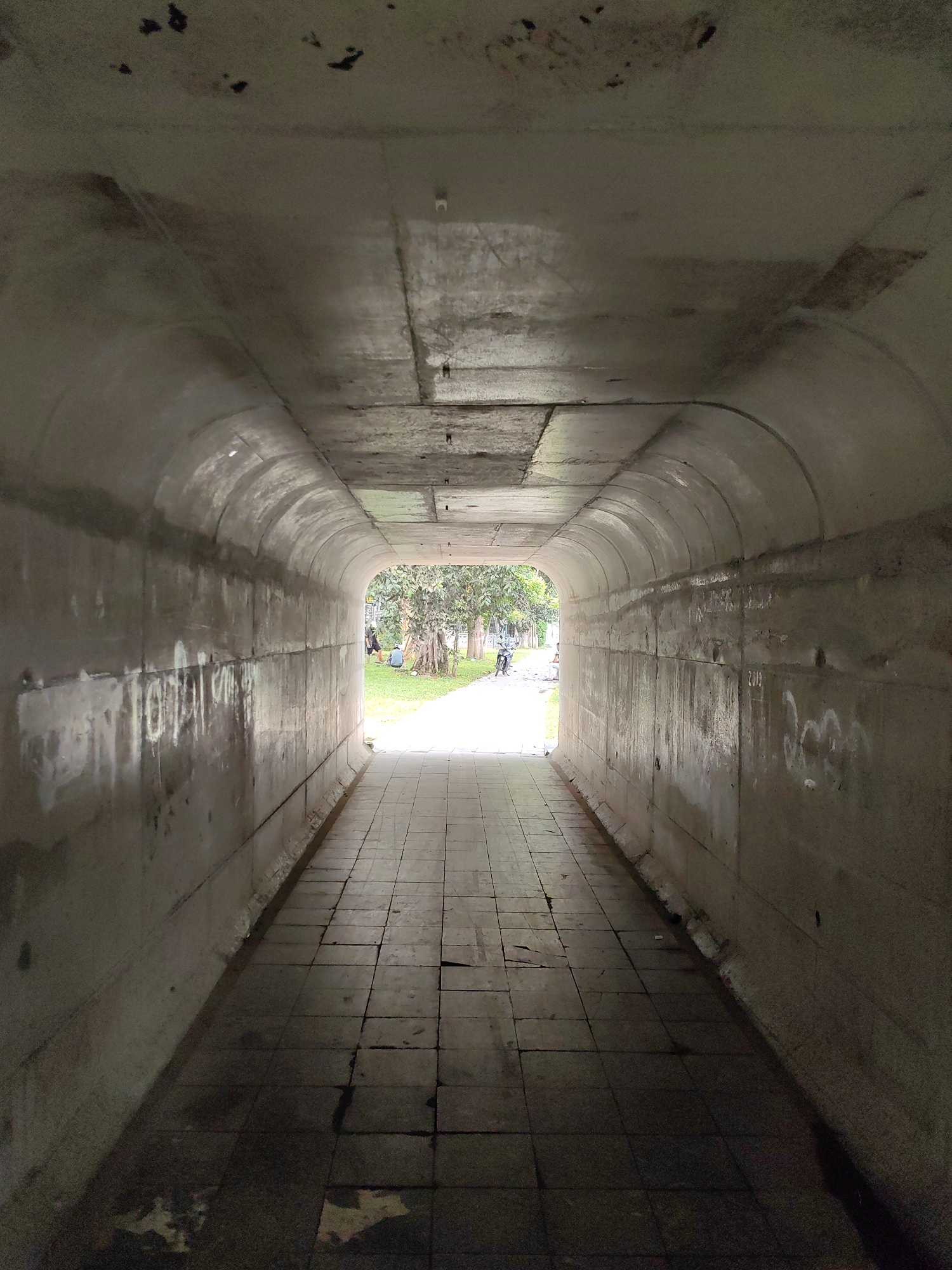 A tunnel in Hà Giang, Vietnam, constructed of concrete and lined with square paving stones, leads to an opening revealing a park with green grass and trees. At the entrance, two individuals are visible, one sitting on the grass and another standing nearby. A scooter is parked on the pathway outside the tunnel. The interior of the tunnel shows signs of weathering and stains, with graffiti scrawled on the walls. The ceiling is a combination of flat concrete slabs and arched segments. The floor of the tunnel is damp.
