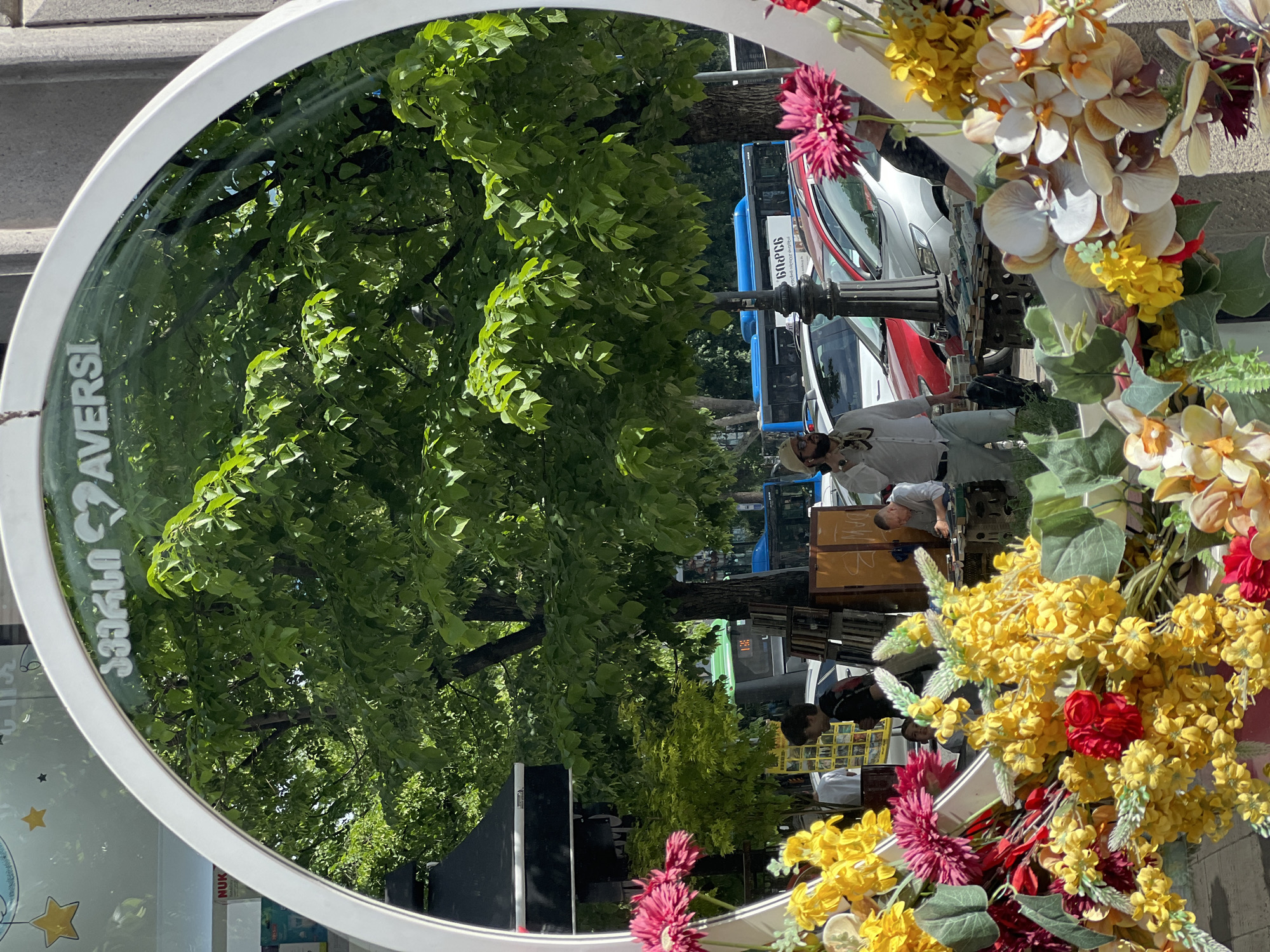 A round mirror decorated with flowers reflects a Tbilisi, Georgia street scene. Lush green trees dominate the reflection. Various vehicles including cars and a blue and white bus are also visible. Several people are reflected in the mirror including a man with a beard wearing a white shirt and headdress, and another person wearing a black shirt. The surrounding environment appears to be a bustling city street.
