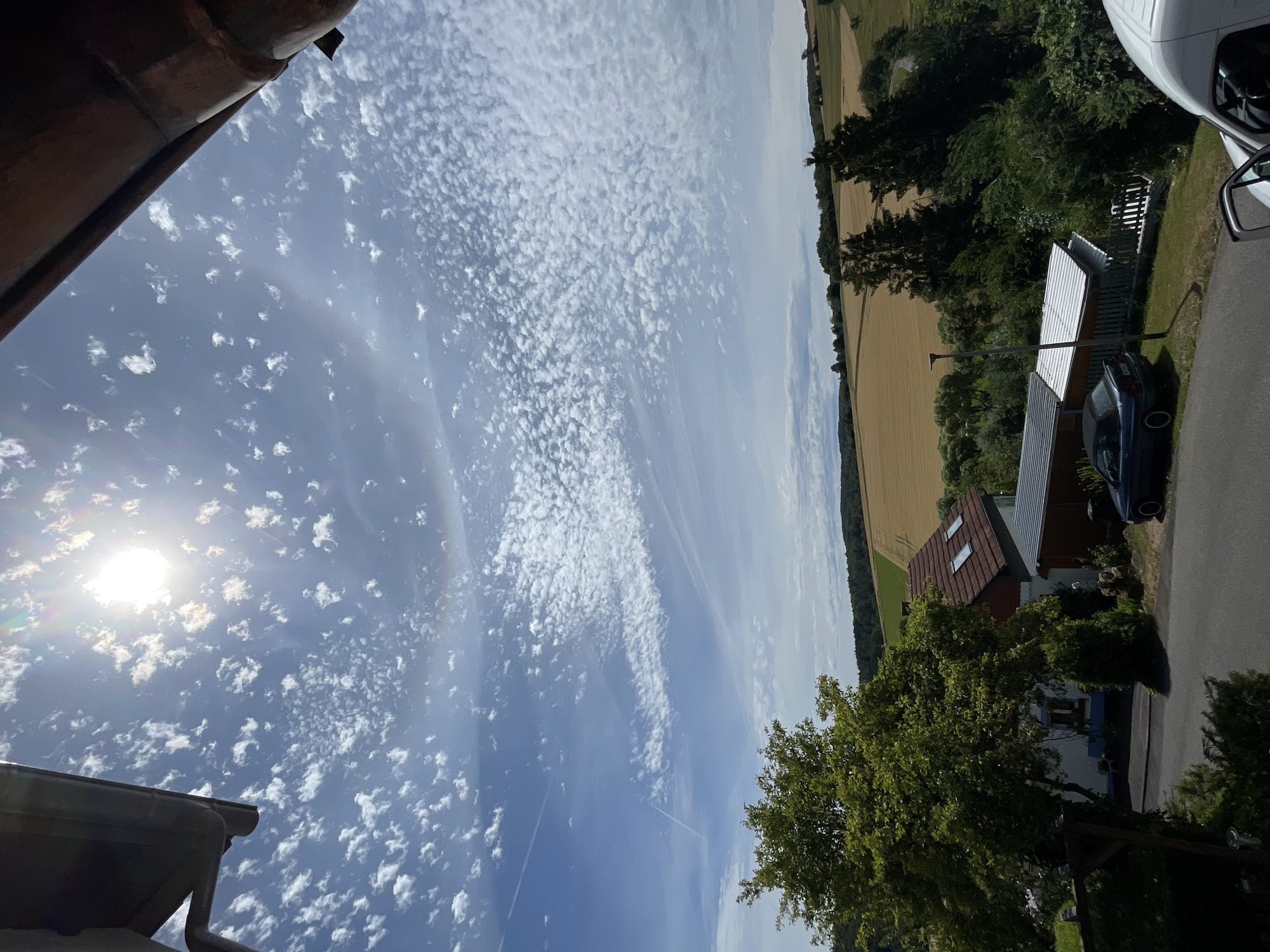 The image displays a rural scene in Schwarzenborn, Germany, on a bright, sunny day with scattered clouds. The sky dominates the upper portion of the frame, featuring a sun that appears to be slightly obscured by thin clouds, creating a subtle halo effect. Numerous small, puffy white clouds are scattered across the blue sky, with some appearing in a wave-like pattern.
The mid-ground reveals a landscape of rolling hills. A large field of golden wheat or similar crop is visible, with green trees and foliage interspersed. A small residential building with a red tile roof and two dormer windows is situated in the lower left of the mid-ground, partially obscured by a large, leafy green tree. Adjacent to the house is a long, narrow structure with a corrugated metal roof.
In the foreground, a grey asphalt road runs horizontally across the bottom of the image. A dark blue convertible car is parked on the side of the road. A white vehicle, possibly a van, is partially visible in the lower right corner. A street lamp stands near the parked car and the building. The overall impression is one of a peaceful, rural setting under a clear, partly cloudy sky.
