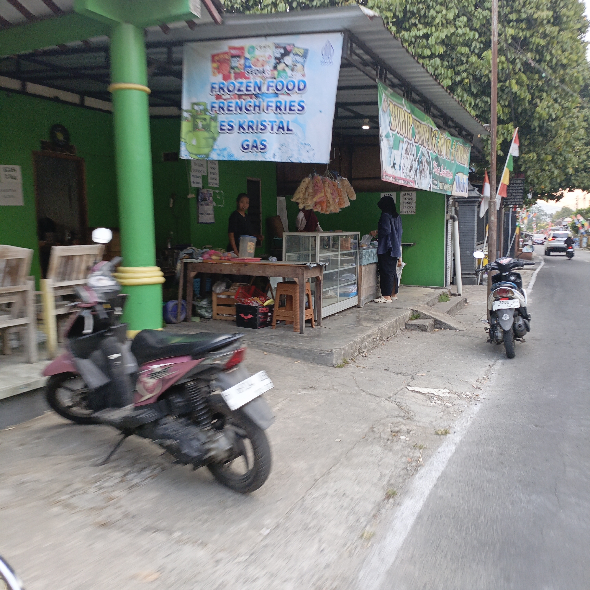 The image captures a street scene in Salatiga, Indonesia, in front of a small food stall. The stall has a green facade and a corrugated metal roof, under which a banner advertises "FROZEN FOOD FRENCH FRIES ES KRISTAL GAS." Several people are visible: a woman in a dark top stands behind a wooden table displaying goods, another person wearing a dark hijab is partially visible near a glass display case, and a child in white stands to the right of the display case.
Two motorcycles are parked on the sidewalk in front of the stall. The one in the foreground is a dark pink scooter, with its kickstand down. Another scooter, primarily white and black, is parked further down the street to the right. The street is paved with asphalt, and the sidewalk is concrete, showing some cracks and wear.
A tall, green cylindrical pole with yellow rings is a prominent feature near the stall. Beyond the stall, the street continues with some traffic, including a white car and another person on a motorcycle in the distance. Trees with dense green foliage line the right side of the street, and Indonesian flags are visible on poles. The overall impression is a bustling roadside area, likely a commercial strip in an Indonesian city.