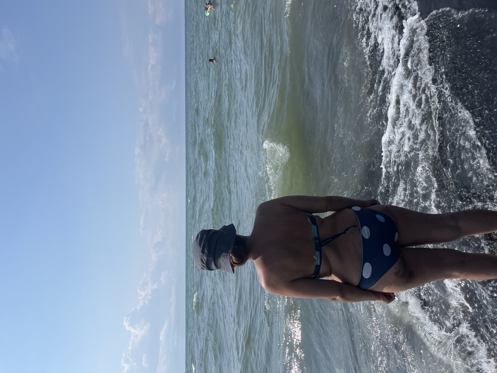 The image depicts a woman from behind, wearing a dark bucket hat and a blue and white polka-dot bikini, standing at the edge of the Black Sea in Ureki, Georgia. The water is choppy with small waves lapping at the dark, pebbly shore. Several people can be seen swimming in the distance. The sky is clear blue with scattered white clouds. The sun is shining brightly, reflecting off the water.