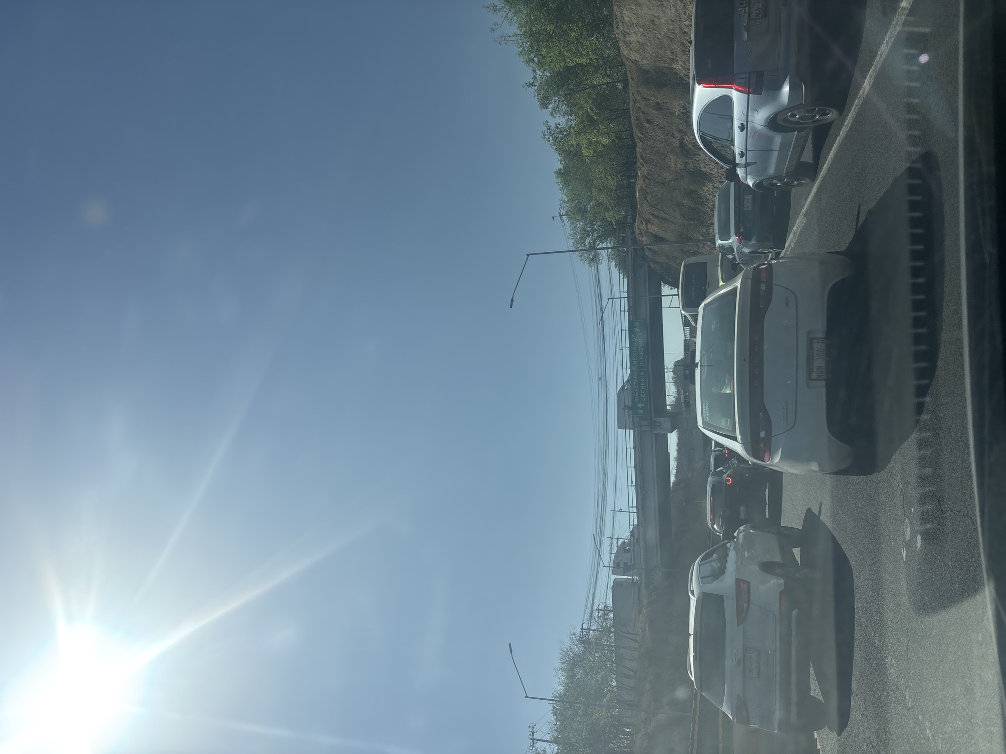 The image depicts a scene of heavy traffic on a multi-lane highway or major road in Cuautitlán Izcalli, Mexico, under a bright, clear blue sky. The photograph is taken from inside a vehicle, likely through a side window, looking forward along the queue of cars. A strong sunburst with prominent lens flare emanates from the upper left corner of the frame, indicating a sunny, clear day, likely around midday.
A line of vehicles is visible, including several white and silver SUVs and minivans, and darker sedans, all facing forward and appearing to be either stopped or moving very slowly in a traffic jam. The asphalt road surface shows visible lane markings.
To the right of the road, a steep, natural embankment covered with dirt, rocks, and green trees rises. Streetlights and overhead utility lines are visible along the route.
Further down the road, a large green overhead highway sign spans across the lanes. Visible white text on the sign indicates directions to local and regional destinations: "QUERETARO", "ATIZAPAN", "LECHERIA", and "TLALNEPANTLA". These place names are consistent with the provided location context of Cuautitlán Izcalli in Mexico. No people are explicitly visible in the frame, but their presence is implied within the vehicles. The scene captures the common experience of urban highway congestion.