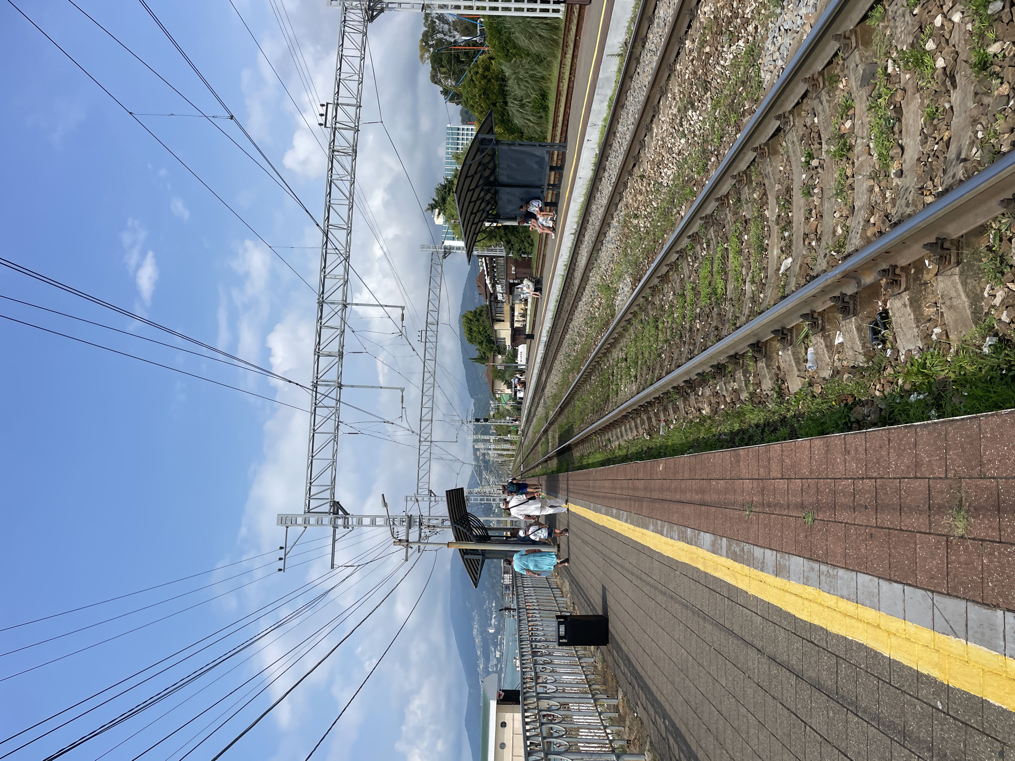This is a photograph taken at a train station in Sochi, Russia, likely near the coast. The scene depicts a train platform with multiple sets of train tracks running parallel. On the left side of the image is a paved platform with a yellow safety line, and further left, a metal railing overlooks a body of water, possibly the Black Sea, with a mountainous coastline in the distance. A black trash bin is visible on the platform. Several people are walking on the platform, including a woman in a blue dress, a child, and two adults in white clothing. Further down the platform, there are covered waiting areas with people seated. Overhead, a complex network of electrical wires and supports for an overhead catenary system for the trains is prominent against a blue sky with scattered clouds. The train tracks are surrounded by gravel and some sparse vegetation. The overall impression is a sunny day at a coastal train station.