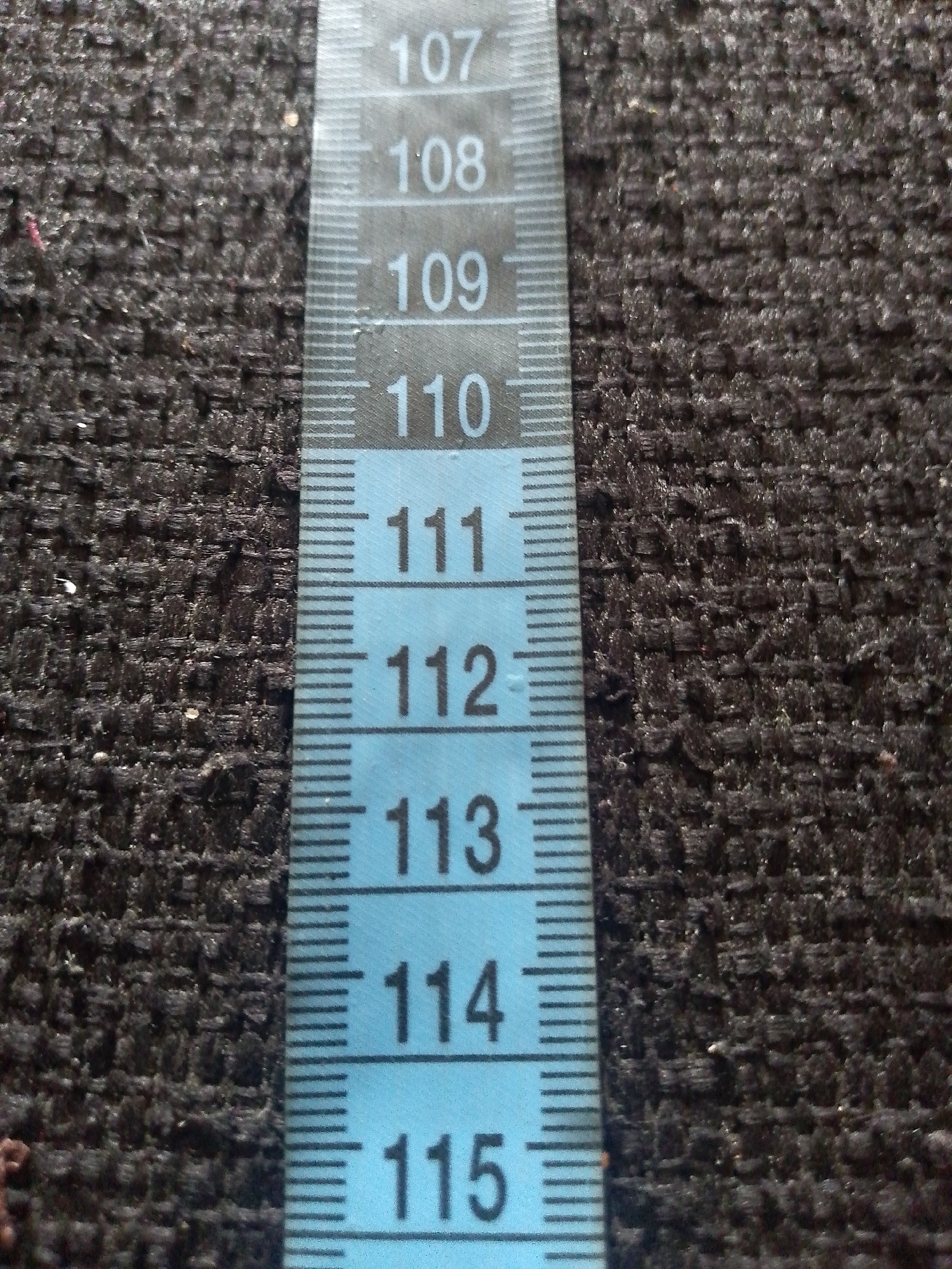 A close-up, eye-level shot shows a blue fabric material with a measuring tape overlaid vertically. The measuring tape has black numbers marking increments, starting from 107 at the top and going down to 115 at the bottom. The fabric has a textured, woven appearance. The lighting appears to be natural, casting subtle shadows on the fabric's surface. No people or animals are visible in the frame. The scene does not provide any information or context related to the city of Jauro Yinu, Nigeria, or any specific urban environment.