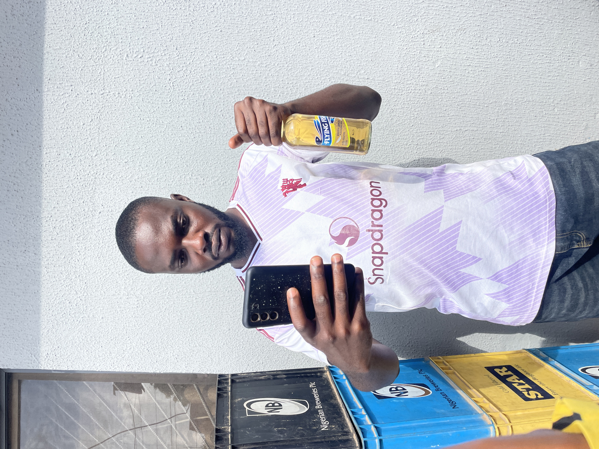 A Black man stands outdoors in front of a textured white wall, possibly in Jos, Nigeria, holding a smartphone in his left hand and a clear glass bottle of Flying Fish Zesty Lemon beverage in his right hand. He is wearing a white and purple athletic jersey with a red crest and the word "Snapdragon" emblazoned across the chest. The bottle, partially filled with a yellow liquid, has a label with "Flying Fish" and "Zesty Lemon" prominently displayed. Behind him to the left, stacks of black and blue plastic crates are visible, with "NB" and "Nigerian Breweries Plc" markings on the black crates, and yellow crates with "STAR" branding. The lighting suggests it is daytime, with sunlight casting a shadow on the wall.
