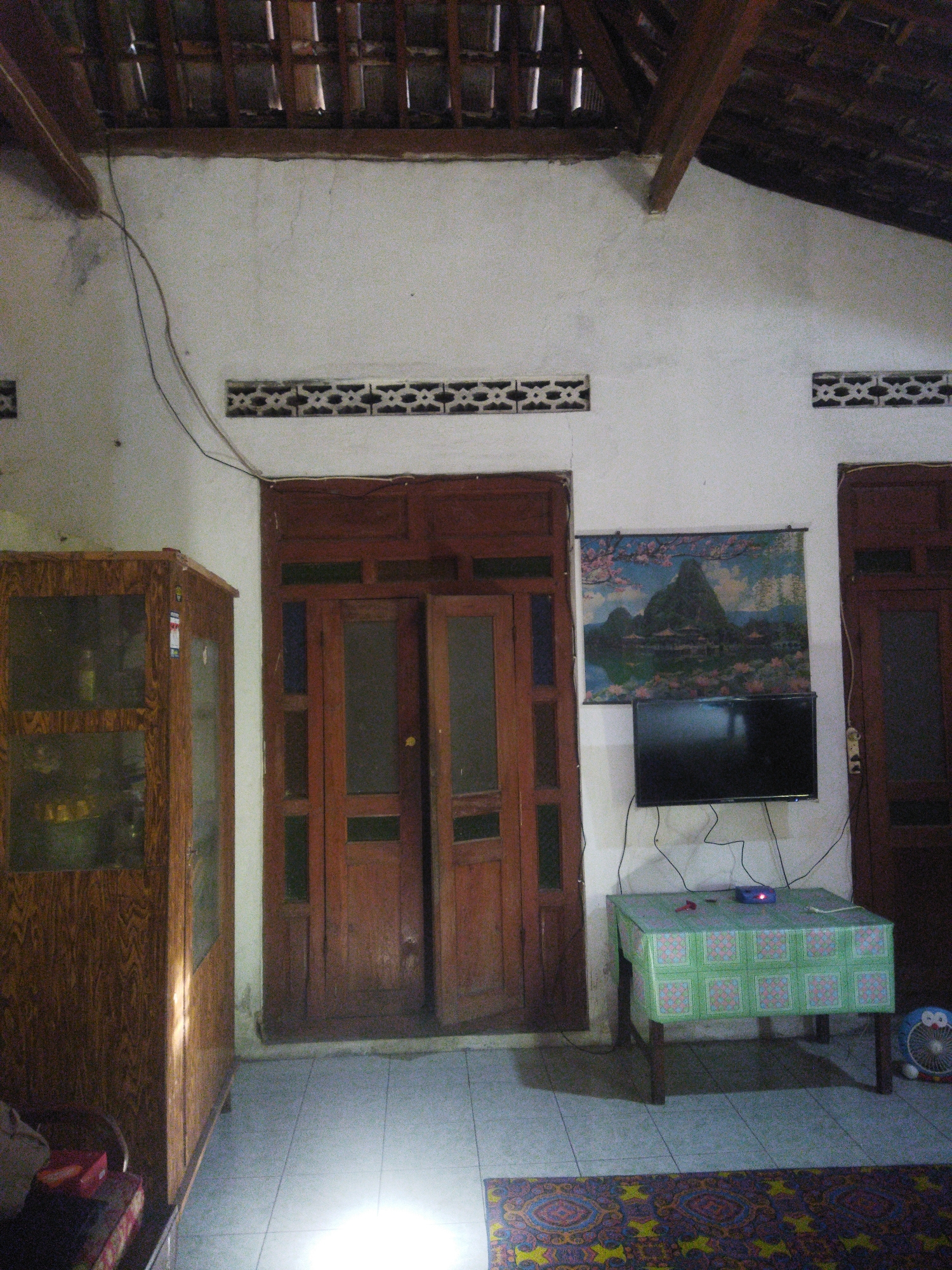 A low-angle, indoor shot shows a living room in Juwiring, Indonesia. The room has a tiled floor and a pitched, wooden ceiling with exposed beams. The walls are white. To the left is a wooden display cabinet with glass doors containing various items. In the center are double wooden doors. To the right of the doors is a wall tapestry depicting a mountainous landscape with a body of water and trees. Below the tapestry is a flat-screen television. In front of the television is a small table covered with a green and white patterned tablecloth. A blue and white fan is visible to the far right. The scene is lit by an overhead light source causing a bright reflection on the floor.