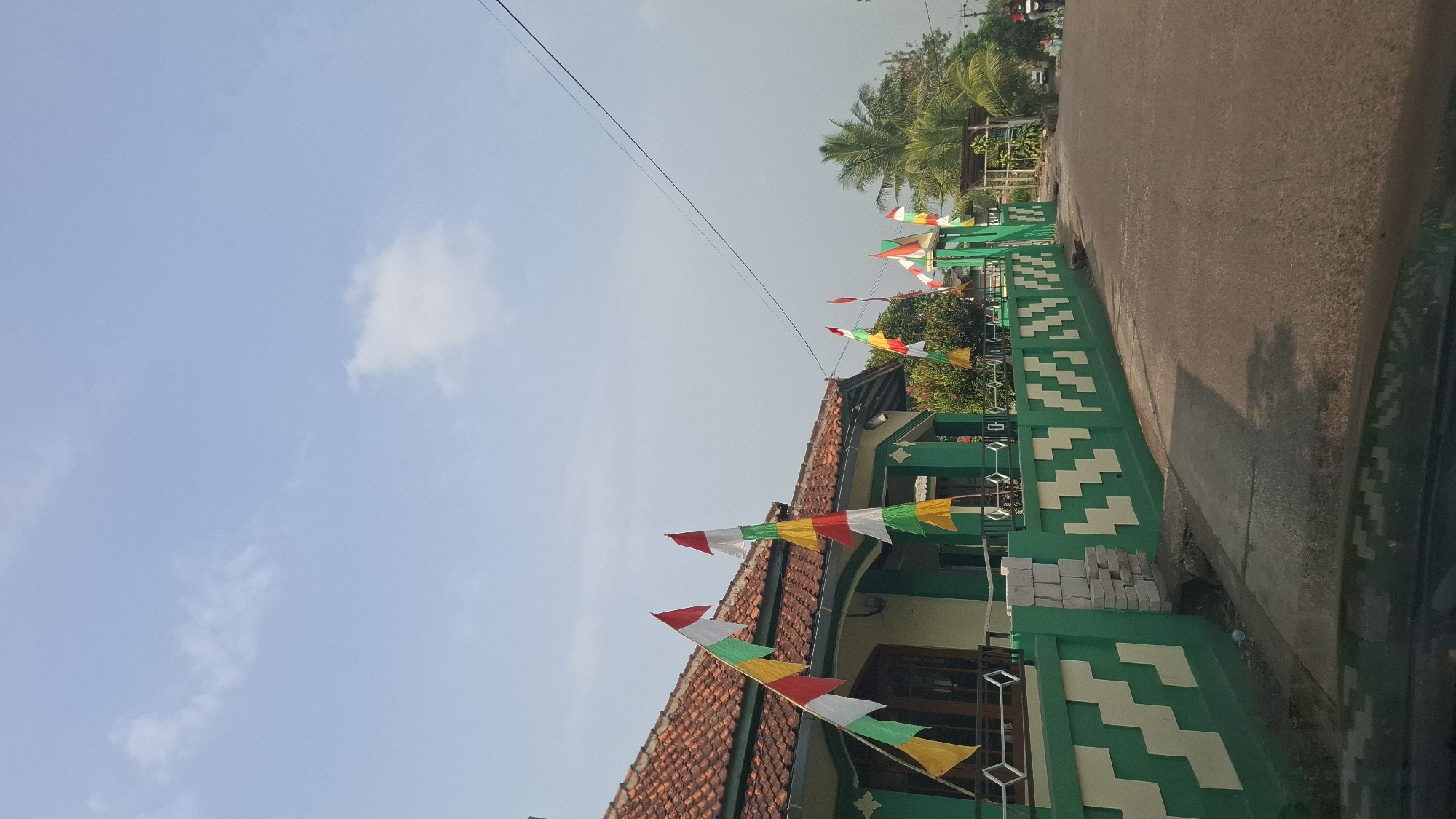 This is a photograph taken on a sunny day in Cibinong Hilir, Indonesia, focusing on a residential property and its surroundings. The foreground shows the roof of a vehicle, indicating the photo was taken from inside a car. The middle ground features a green and white patterned wall with a residential building behind it, characterized by a red tiled roof and green trim. Several Indonesian flags, with red and white stripes, are displayed prominently along the wall and near the house. Beyond the property, a paved road runs alongside, with trees, including palm trees, and other vegetation visible. In the distance, a person on a motorcycle is seen on the road. The sky above is clear with scattered white clouds.