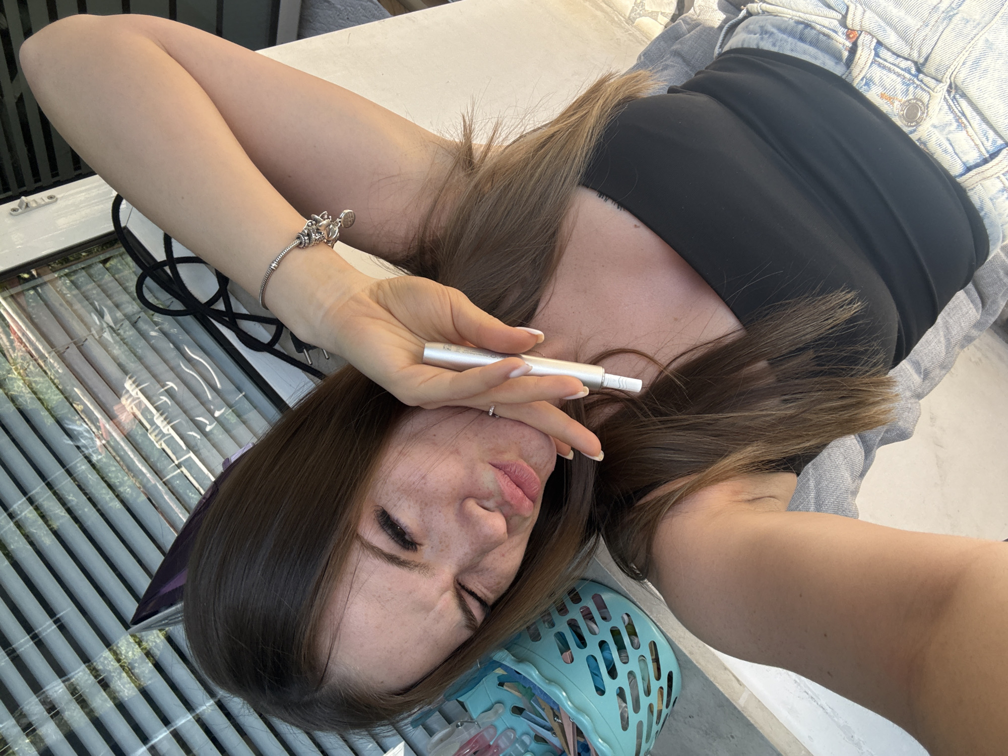 A young woman is lying down, seemingly on an outdoor surface in what appears to be a residential or semi-public space. She is holding a small, cylindrical object in her hand near her face. She is wearing a black tank top, a denim jacket, and a bracelet on her left wrist. Her hair is brown and shoulder-length. Her face shows visible freckles. To the left, there's a window with blinds. A blue plastic basket with various items inside is positioned near her head. There is no visible signage to pinpoint the exact location in Plovdiv, Bulgaria.