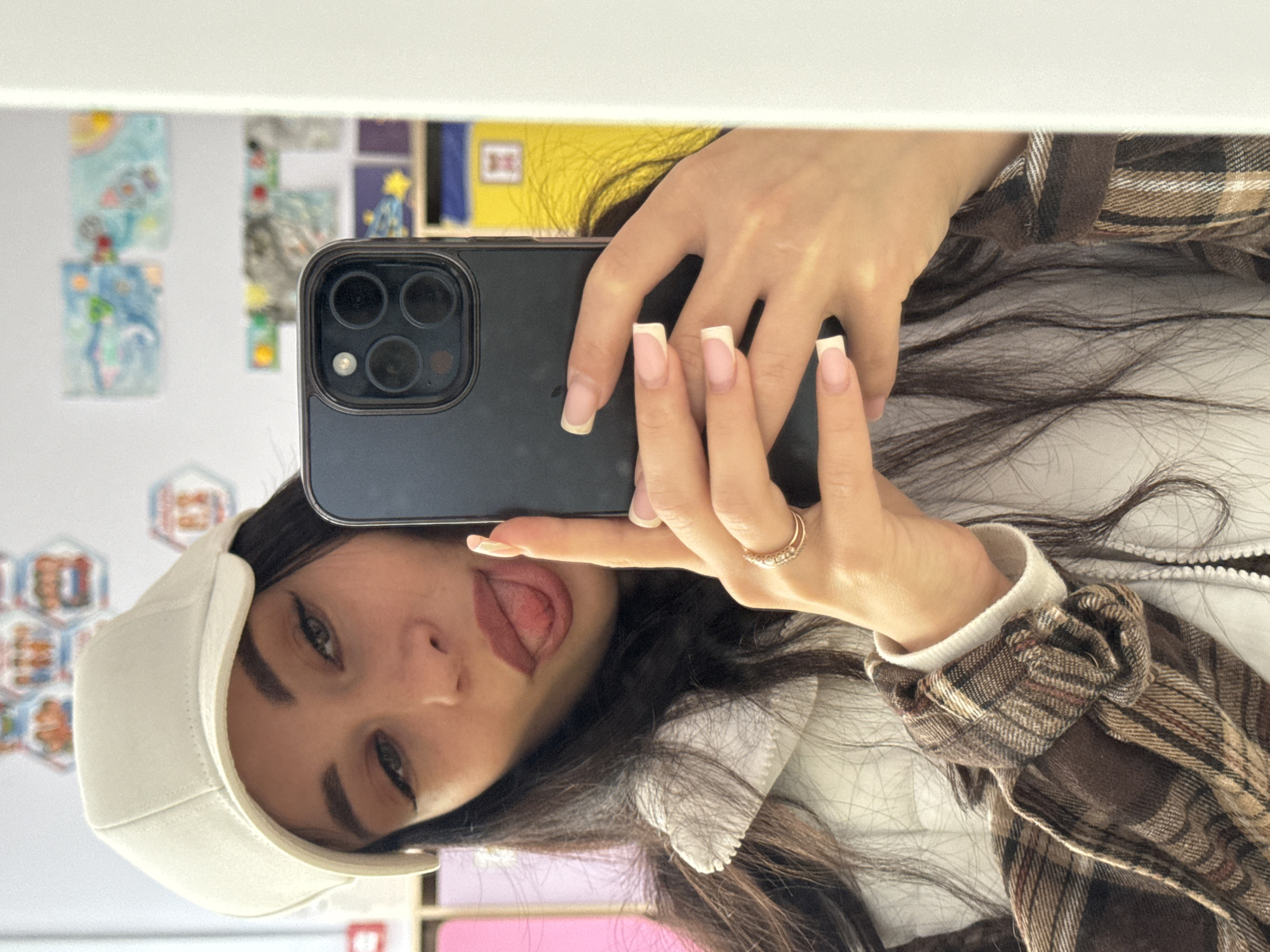 A young woman is taking a selfie in what appears to be an indoor setting. She is holding a dark-colored smartphone with a triple-lens camera in her right hand. Her left hand is also visible, partially obscuring the phone. She is sticking her tongue out playfully. The woman is wearing a white cap, a white vest with a zipper, and a brown plaid shirt. She has long dark hair and wears a ring on one of her fingers. The background includes artwork or pictures on the wall, along with other indistinct objects. The location in Chaikovsky, Russia is not discernible from the image.