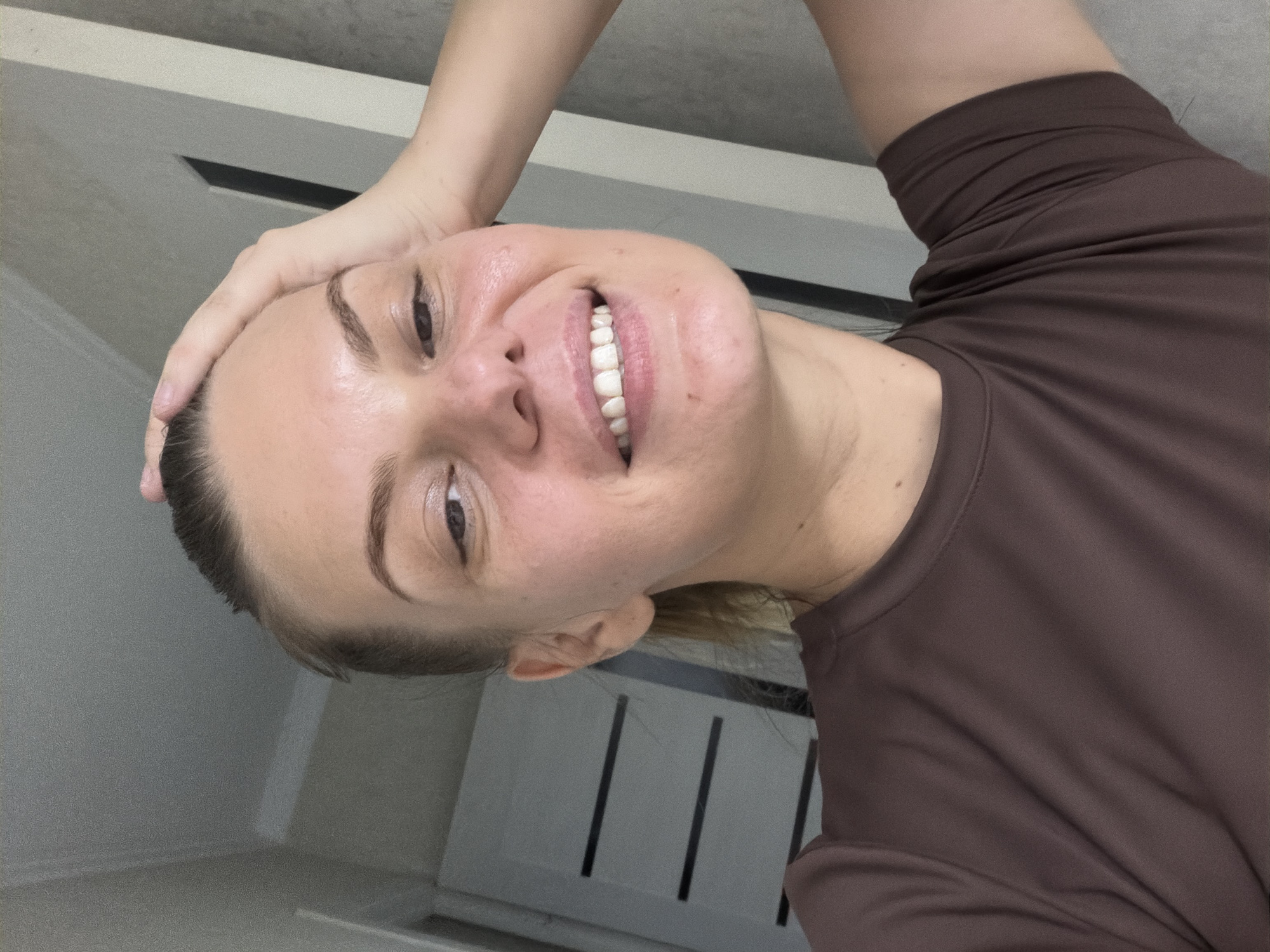 A woman is taking a selfie indoors. She has her right hand on top of her head and is smiling. She is wearing a brown t-shirt. The background shows a doorway and a wall. The lighting suggests it is daytime. There is no discernible information within the image to place the location in Chirchik, Uzbekistan.