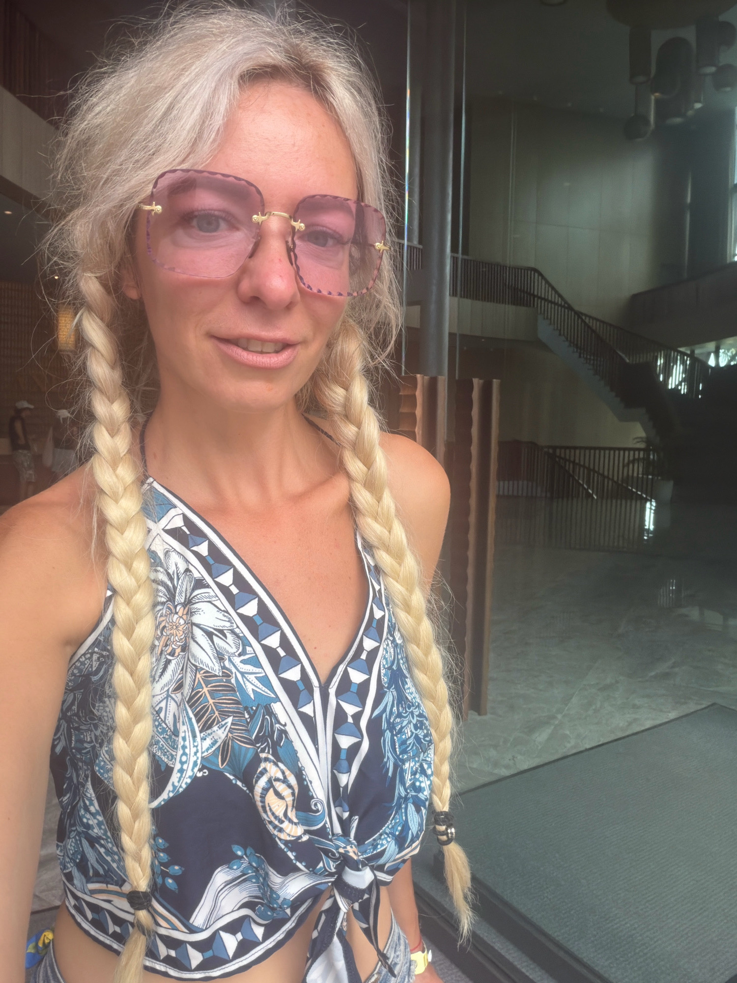 A blonde woman with blue eyes and pink-tinted glasses stands in what appears to be the lobby of a hotel in Cam Lâm, Vietnam. She has her hair in two long braids and is wearing a blue and white patterned halter top. Behind her, a dark, modern staircase with railings is visible, suggesting an indoor public space with multiple levels. The floor appears to be tiled or polished concrete.
