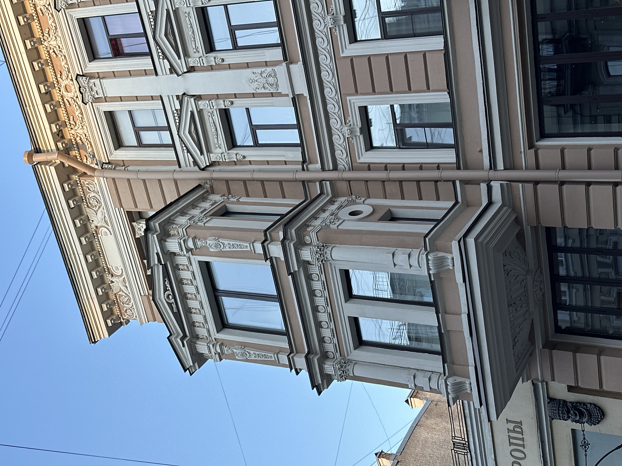 The image shows a section of a building in Saint Petersburg, Russia. The architecture is ornate, featuring decorative moldings, window frames with detailed embellishments, and fluted columns. The building is painted in beige and white tones, characteristic of the city's historical architectural style. The sky is visible in the background. A vertical drainpipe runs along the building's facade.