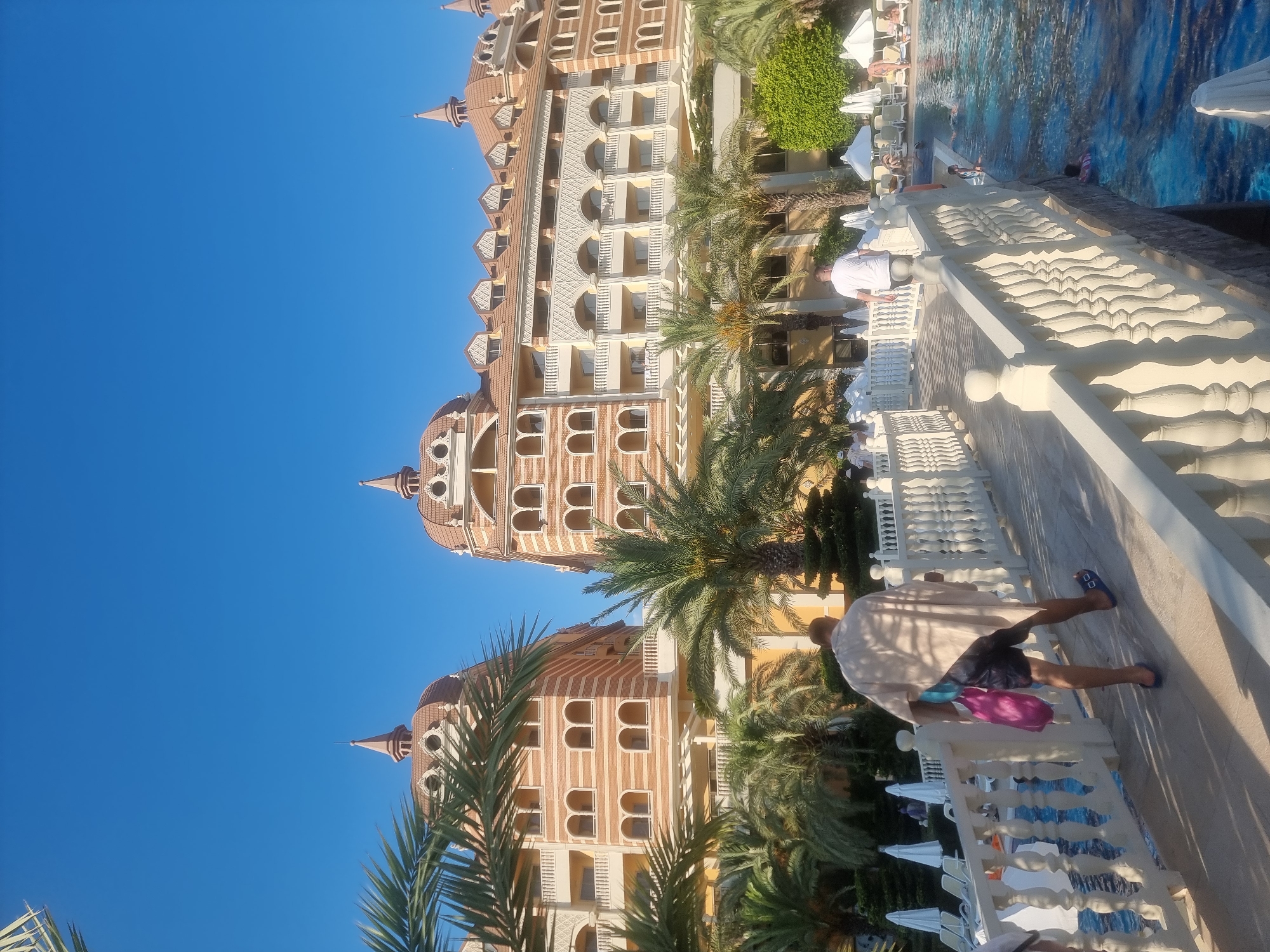 A clear blue sky looms over a resort in Manavgat, Turkey. Two opulent, multi-story buildings with distinctive dome-shaped roofs and ornate facades dominate the background. Palm trees are scattered throughout the scene, adding to the tropical ambiance. In the foreground, a white, ornate bridge spans across a vibrant blue swimming pool. A man in a light-colored robe and dark shorts walks across the bridge, carrying two shopping bags. Other individuals are visible near the pool, some relaxing on lounge chairs and others swimming. The scene conveys a sense of leisure and vacation.
