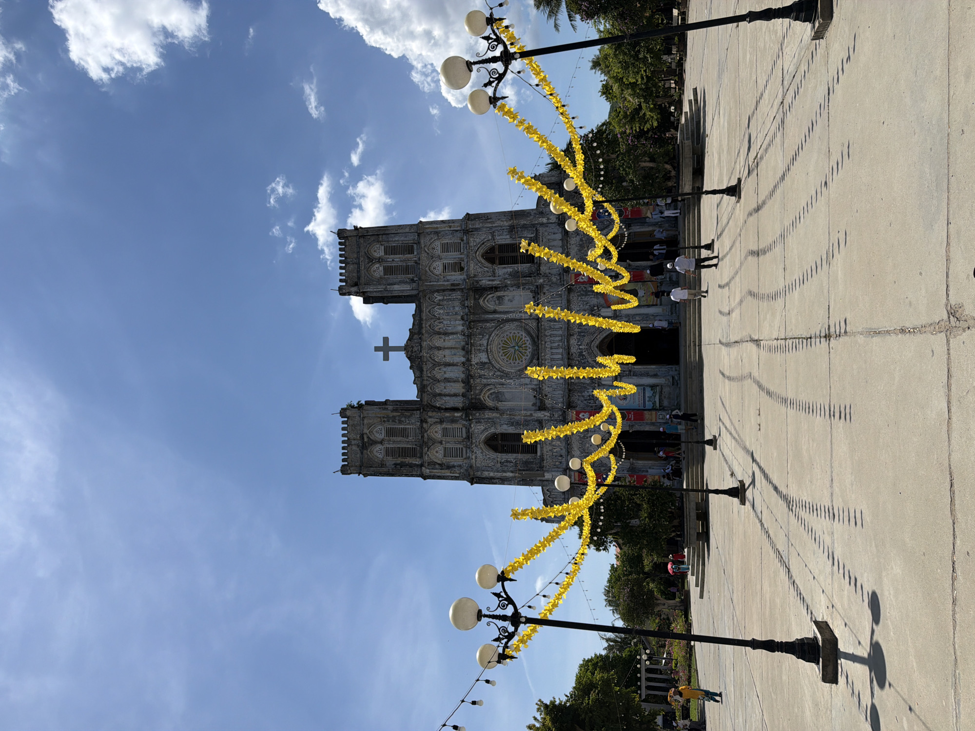 The image depicts a large, ornate church with two bell towers and a prominent cross on its facade. The church is constructed of stone and exhibits intricate architectural details. In front of the church, a paved plaza stretches out, adorned with yellow decorative garlands and strings of lights. Several streetlights with multiple globes stand on either side of the plaza, casting long shadows on the ground. A few individuals are visible in the plaza, some walking towards the church and others standing around. Trees line the perimeter of the plaza, and the sky above is a clear blue with scattered white clouds. This scene is located in Hội Tín, Vietnam, a place known for its religious sites.