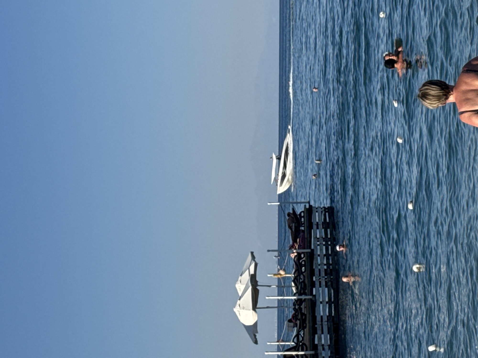 The image captures a sunny day at a beach in Kemer, Turkey. In the foreground, a woman with blonde hair is facing away from the camera, looking out at the sea. The sea is a vibrant blue, with gentle waves. A few white buoys are scattered across the water.
Further out, two people are swimming. One person appears to be closer to the camera, while another pair are further back, possibly talking or embracing.
To the left, a wooden pier extends into the water. On the pier, several empty lounge chairs are set up under two white umbrellas. A few people are visible lounging on the chairs.
In the background, a sleek white speedboat is cutting through the water, leaving a white wake. Beyond the sea and the speedboat, distant mountains are visible against a clear blue sky. The overall scene depicts a tranquil beach resort atmosphere.