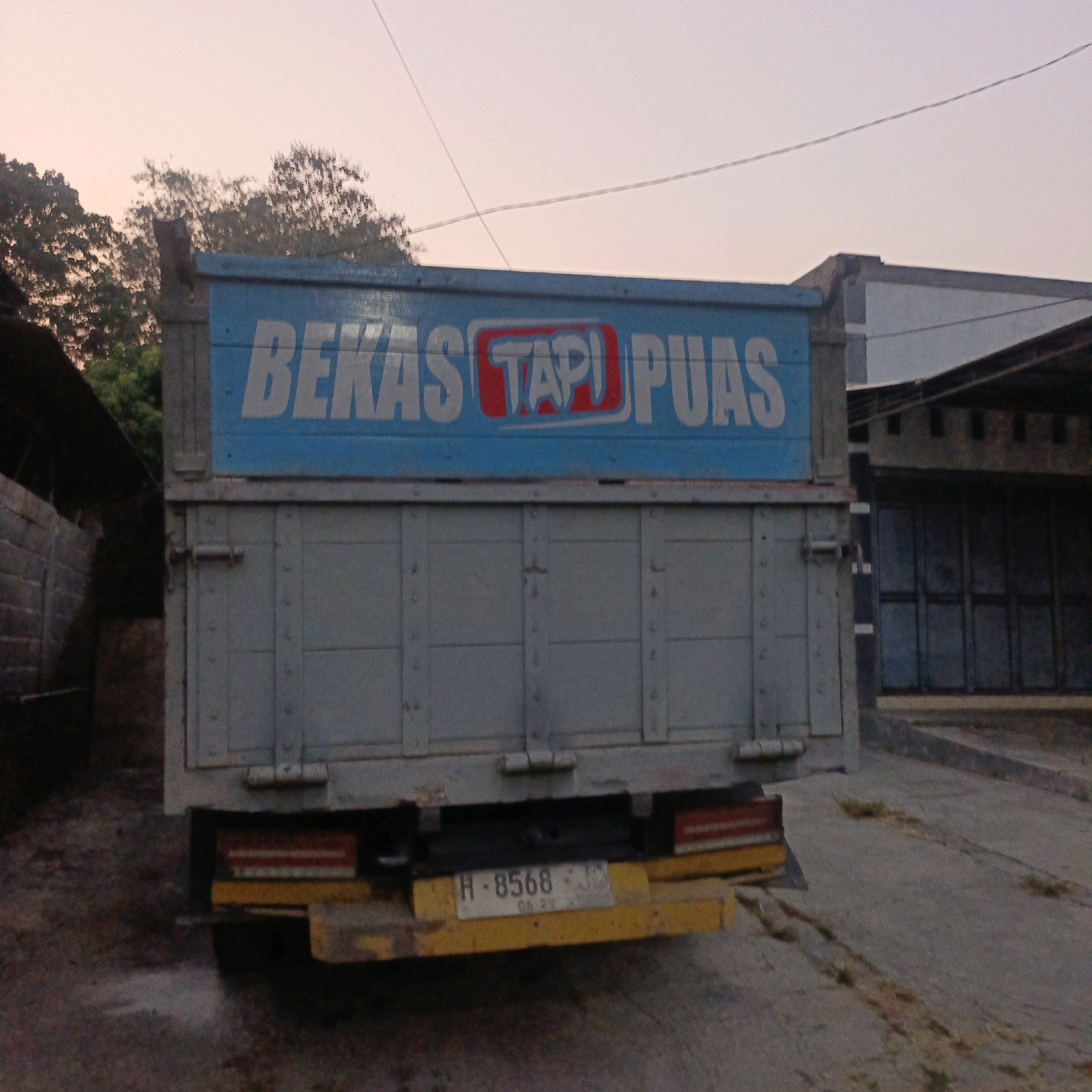 The image shows the rear of a light blue truck with text printed in white on its side. The text reads "BEKAS TAPI PUAS". A red rectangle with white text "TAPI" is highlighted within the larger text. The truck is parked on a concrete surface. In the background, there are trees, a building with a dark facade, and closed metal doors. The sky is a pale pinkish-orange, suggesting dusk or dawn. The truck's license plate is visible and reads "H-8568-JC" with "04 29" below it. The overall scene appears to be in an outdoor urban or semi-urban setting in Salatiga, Indonesia, likely a storage or loading area.