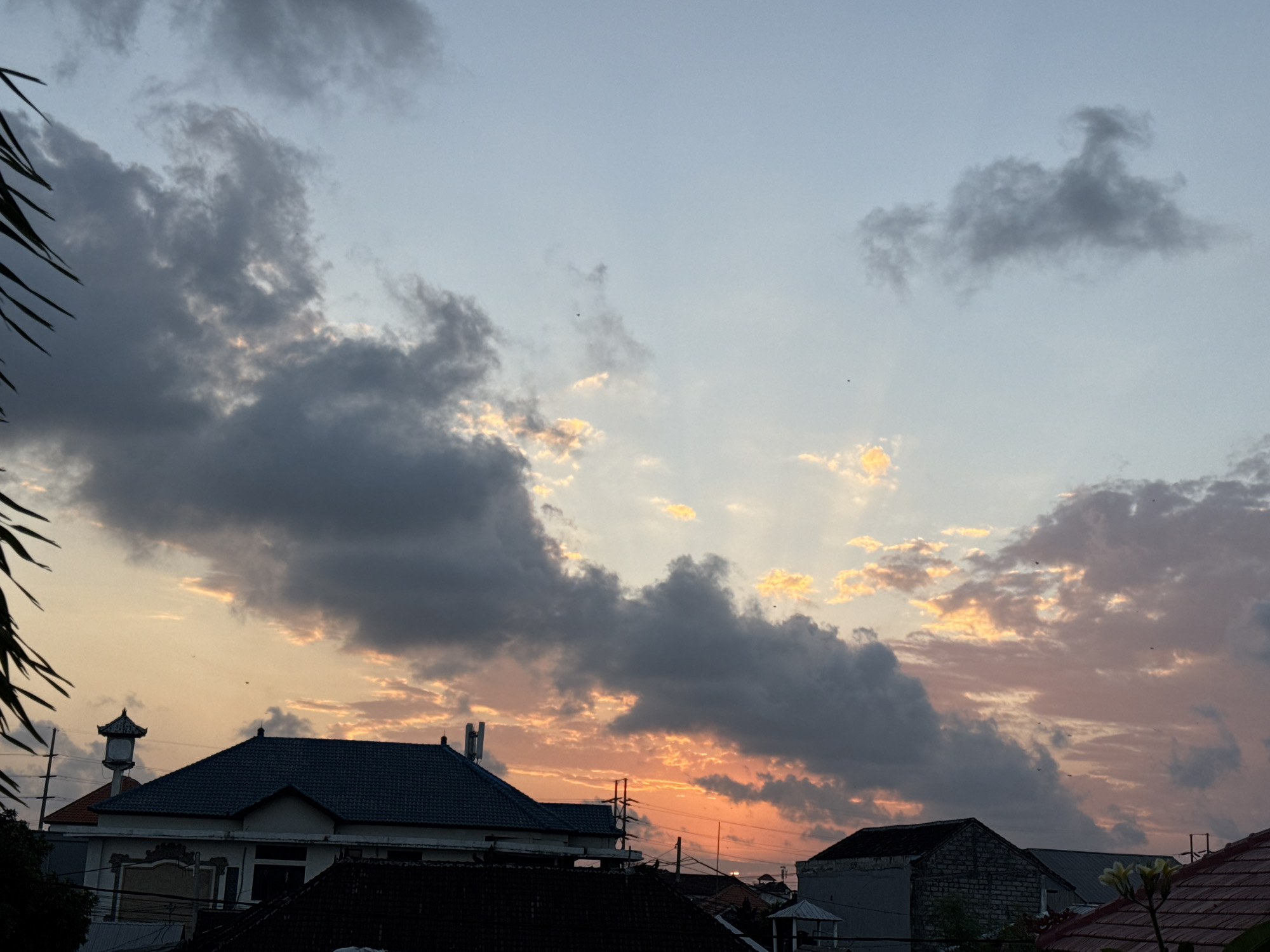 The image depicts a sunset scene over a residential area in Denpasar, Indonesia. Dark gray clouds with tinges of orange and yellow dominate the sky, indicating the setting sun. Silhouetted rooftops of houses, some with dark tiled roofs and others with lighter colored walls, are visible in the foreground. A building with a distinctive tiered roof and a water tank is to the left. In the upper portion of the sky, lighter clouds are scattered against a pale blue and pink gradient. Some thin, dark branches of a palm tree are in the far left of the frame. The overall impression is of a calm evening in an urban environment.