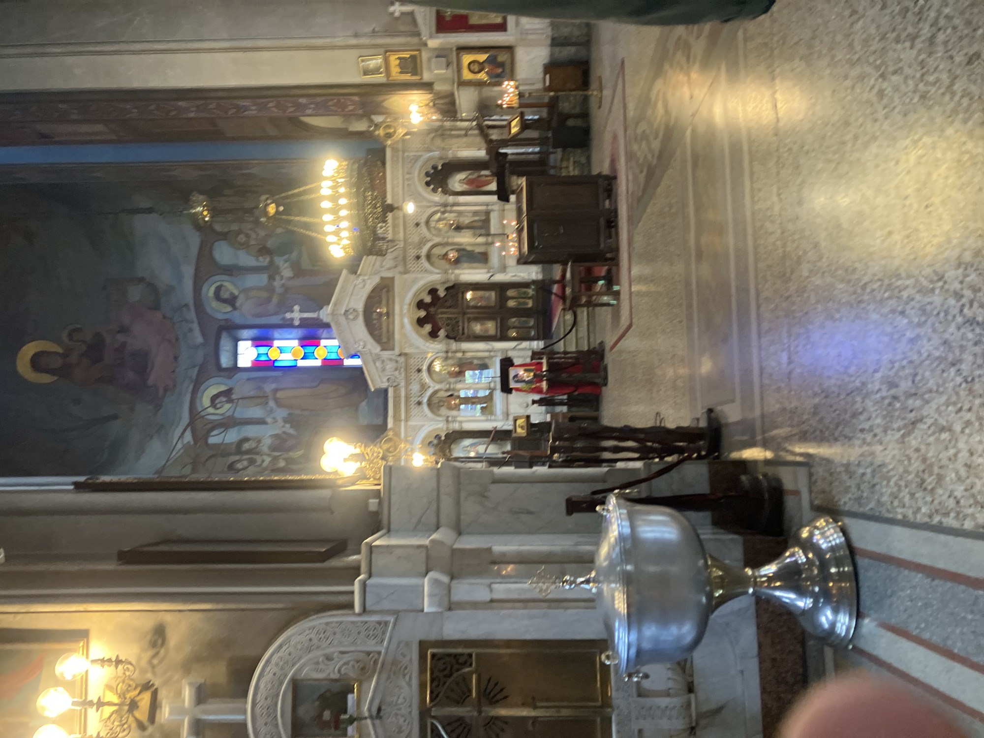 The image depicts the interior of a cathedral in Tbilisi, Georgia. A large, silver baptismal font is prominently displayed in the foreground. The altar area is visible, featuring a decorative iconostasis adorned with paintings and lit by chandeliers. Stained glass windows provide colorful illumination. The walls are decorated with frescoes. The floor is covered in marble.