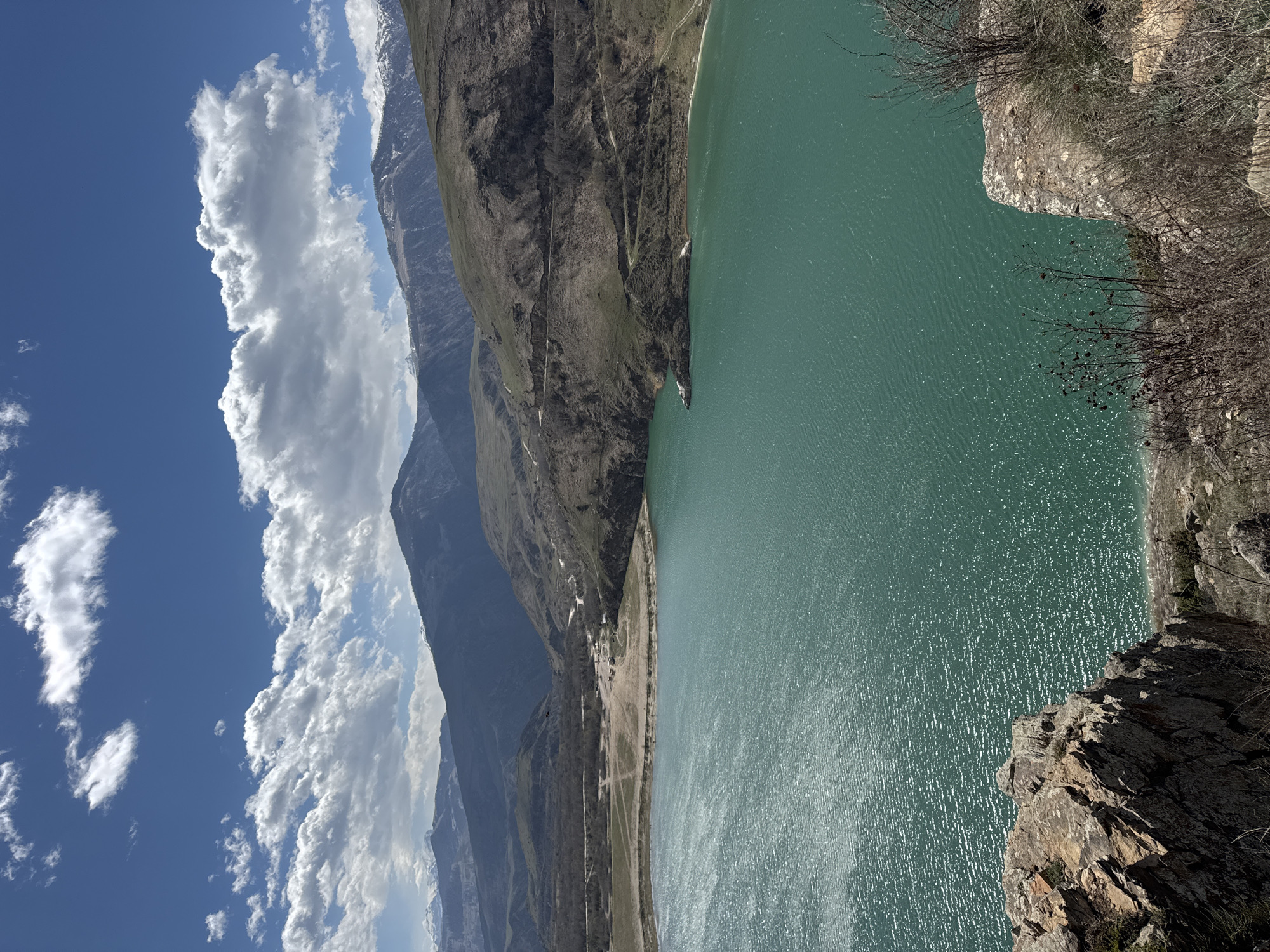 A vertically oriented landscape photograph depicts a scenic view from a height. In the foreground, the edge of a rocky cliff is visible, partially obscured by bare branches. Beyond the cliff, a large body of water stretches out, its surface shimmering with light. On the opposite side of the water, a tall mountain range rises, its slopes covered in various shades of brown and green. Snow is visible on the highest peaks. The sky above is blue, filled with fluffy white clouds that partially obscure the mountain peaks. This is Lake Bylym in Bylym, Russia.