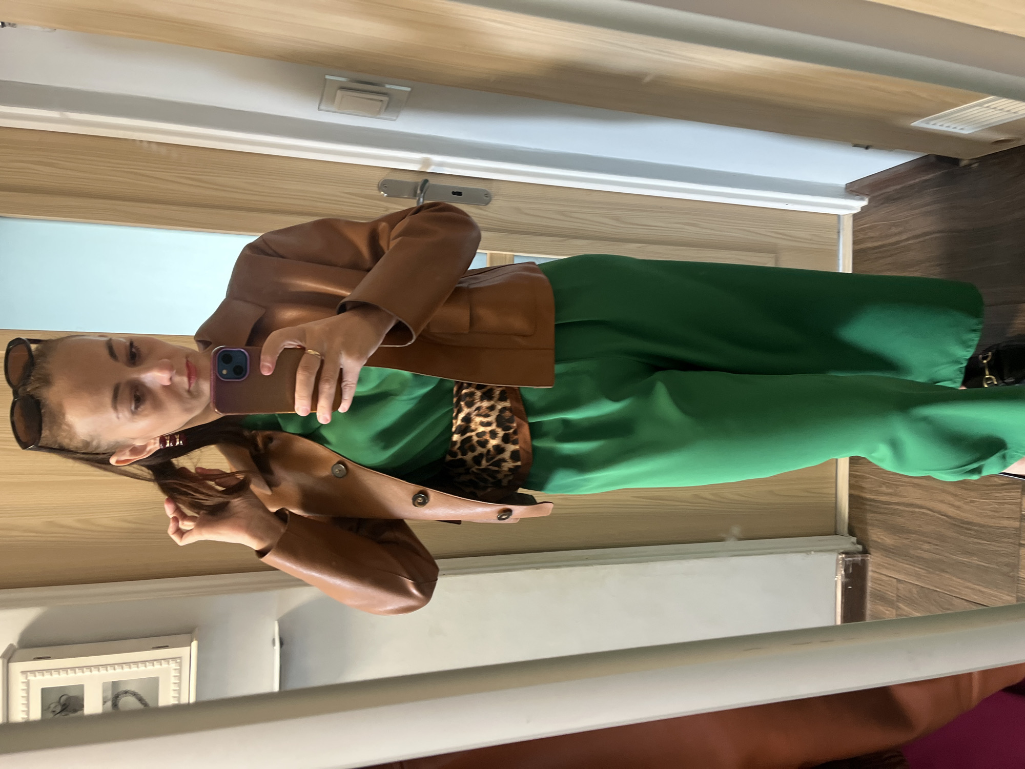 A woman is standing in front of a mirror, taking a selfie. She is wearing a brown leather jacket over a green jumpsuit and a leopard print belt. She has her hair pulled back in a ponytail and is wearing sunglasses on her head. The background appears to be a hallway with wooden doors and a wooden floor. There is also a white cabinet on the left side of the image.