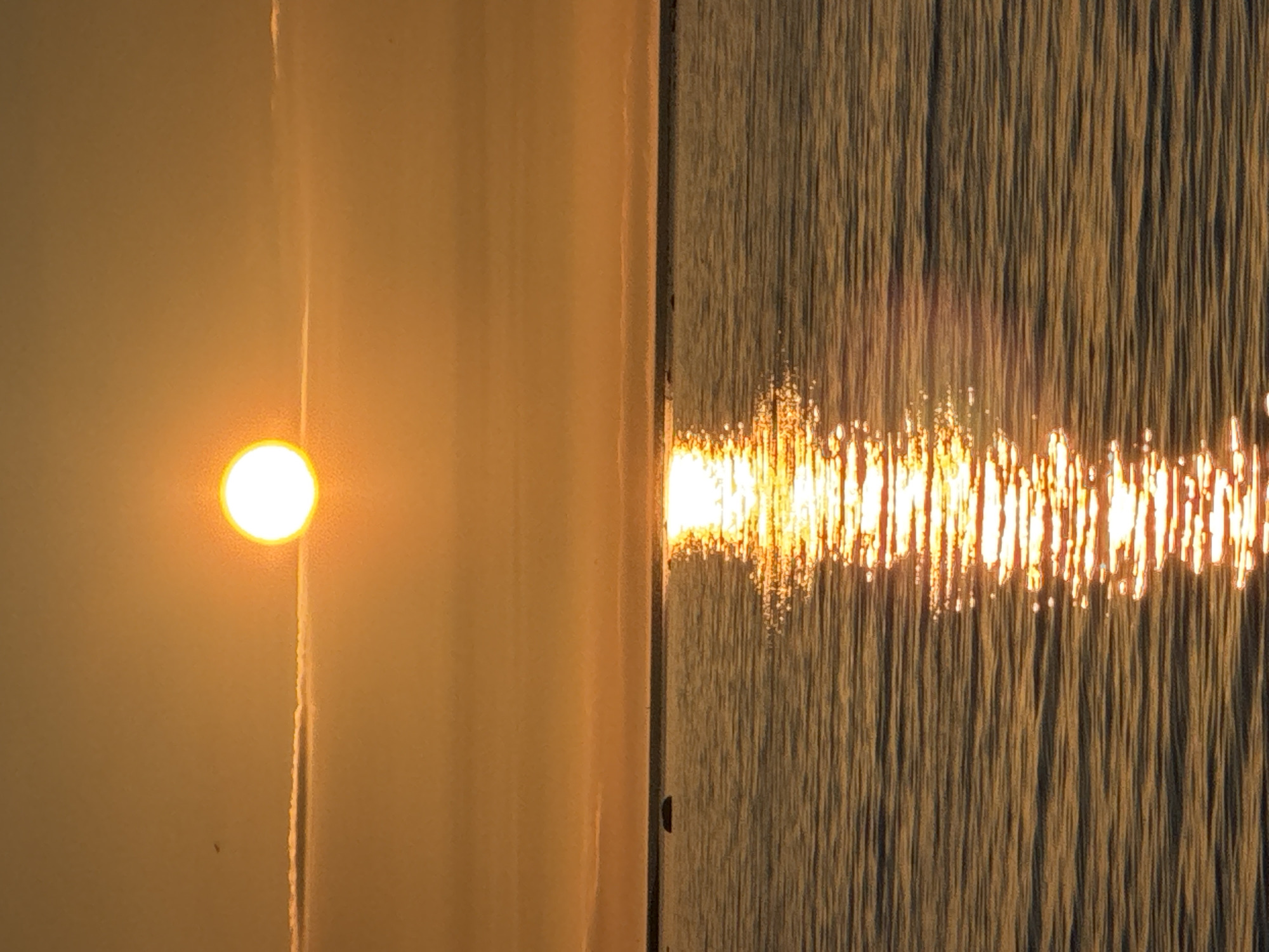 The image depicts a sunrise or sunset over a body of water in Tallinn, Estonia. A luminous white-yellow disc, representing the sun, is positioned high in the frame, slightly left of center, against a sky dominated by warm amber and golden hues. A distinct, thin, horizontal cloud formation stretches across the mid-sky, directly beneath the sun, with fainter, diffuse cloud bands also visible. A tiny, dark speck is present in the upper left quadrant of the sky. The horizon line is sharp and dark, separating the sky from the water. The water surface, reflecting the sky's warm tones, displays ripples and small waves. A prominent, shimmering column of golden-yellow sunlight extends vertically from the horizon towards the bottom of the frame, indicating the sun's direct reflection. Several dark, indistinct objects, consistent with rocks or small islets, break the water's surface along the distant horizon, with one notably larger, rounded object positioned on the left side of the mid-ground water. No discernible people or explicit human actions are present within the scene.