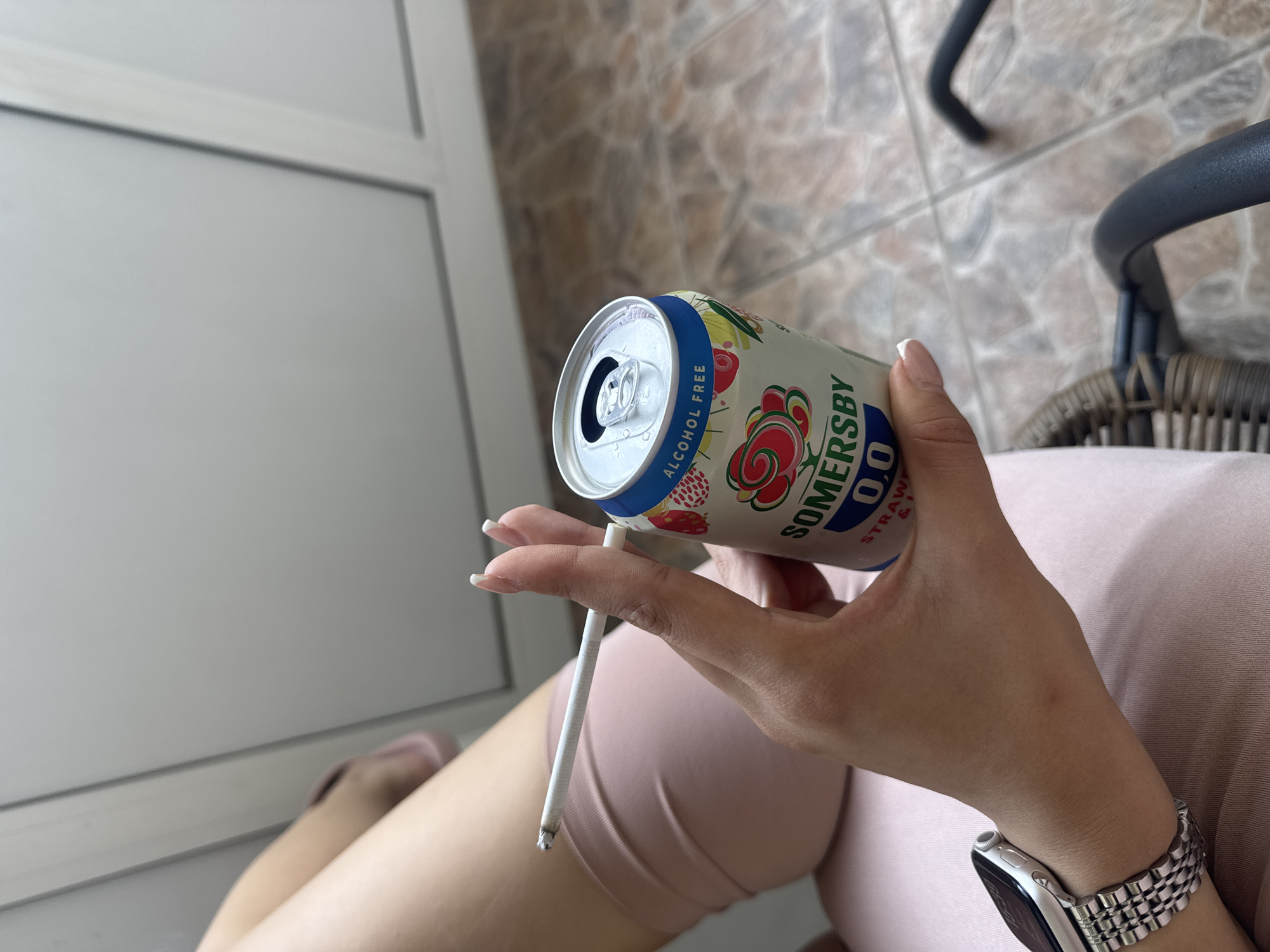 The image captures a close-up, low-angle perspective of a person's hands and upper legs. The primary subject is a hand holding an open can of Somersby 0.0 Alcohol Free Strawberry beverage, with the label "ALCOHOL FREE" clearly visible. The same hand holds a lit cigarette, with a small amount of ash at its tip, between the index and middle fingers. On the person's left wrist, a silver-colored smartwatch with a metal link band is worn.
The person is dressed in light pink or peach-colored clothing, which appears to be soft fabric, possibly a dress or shorts. The setting seems to be an outdoor or semi-outdoor space, such as a balcony or patio in Plovdiv, Bulgaria. The background on the right features a light brown and beige tiled floor and a dark-colored, possibly wicker or rattan, chair. On the left, a white wall or partition with a grey horizontal line adds to the architectural background.
The activity depicted is one of relaxation, as the person is holding a non-alcoholic drink and a cigarette. The lighting suggests it is daytime, with no harsh shadows indicating direct sunlight, implying pleasant weather. No explicit emotions are visible, but the scene suggests a moment of calm.