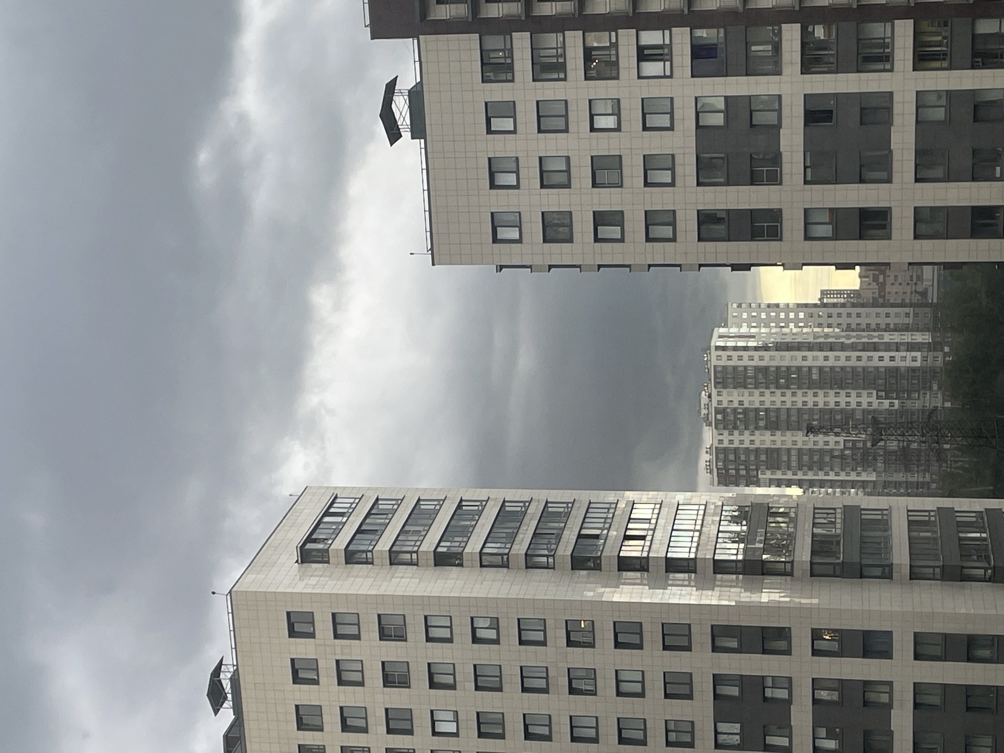The image shows a view of a city skyline with several tall apartment buildings under a cloudy sky. In the foreground, on the left, is a very tall, modern apartment building with a light-colored facade and dark gray accents around the windows. The windows are arranged in a grid pattern. The building on the right is also a tall apartment building, with a similar light-colored facade and dark gray sections. Behind these two buildings, further in the distance, are more apartment buildings, appearing lighter in color and densely packed. A tall, dark metal structure, resembling a transmission tower, is visible between the closer buildings and the distant ones. The sky is heavily overcast with dark, moody clouds, suggesting an impending storm or inclement weather. The overall impression is of a dense urban residential area.