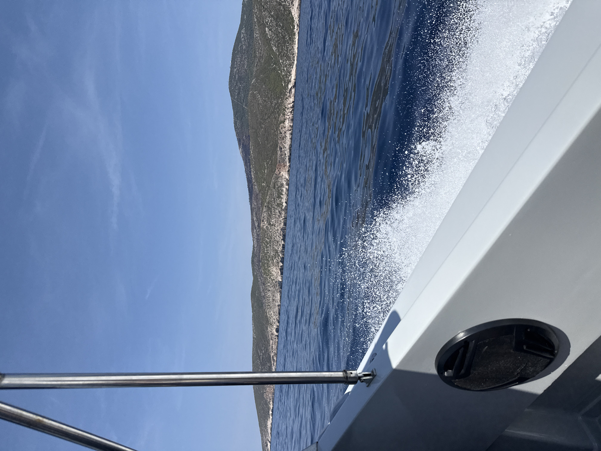 A boat is cutting through the deep blue Adriatic Sea, generating a white spray of foam along its starboard side. In the distance, a green, hilly island with rocky coastal cliffs is visible under a clear blue sky. A metal railing and part of the boat's white hull are in the foreground. The location is near Podhumlje, Croatia.