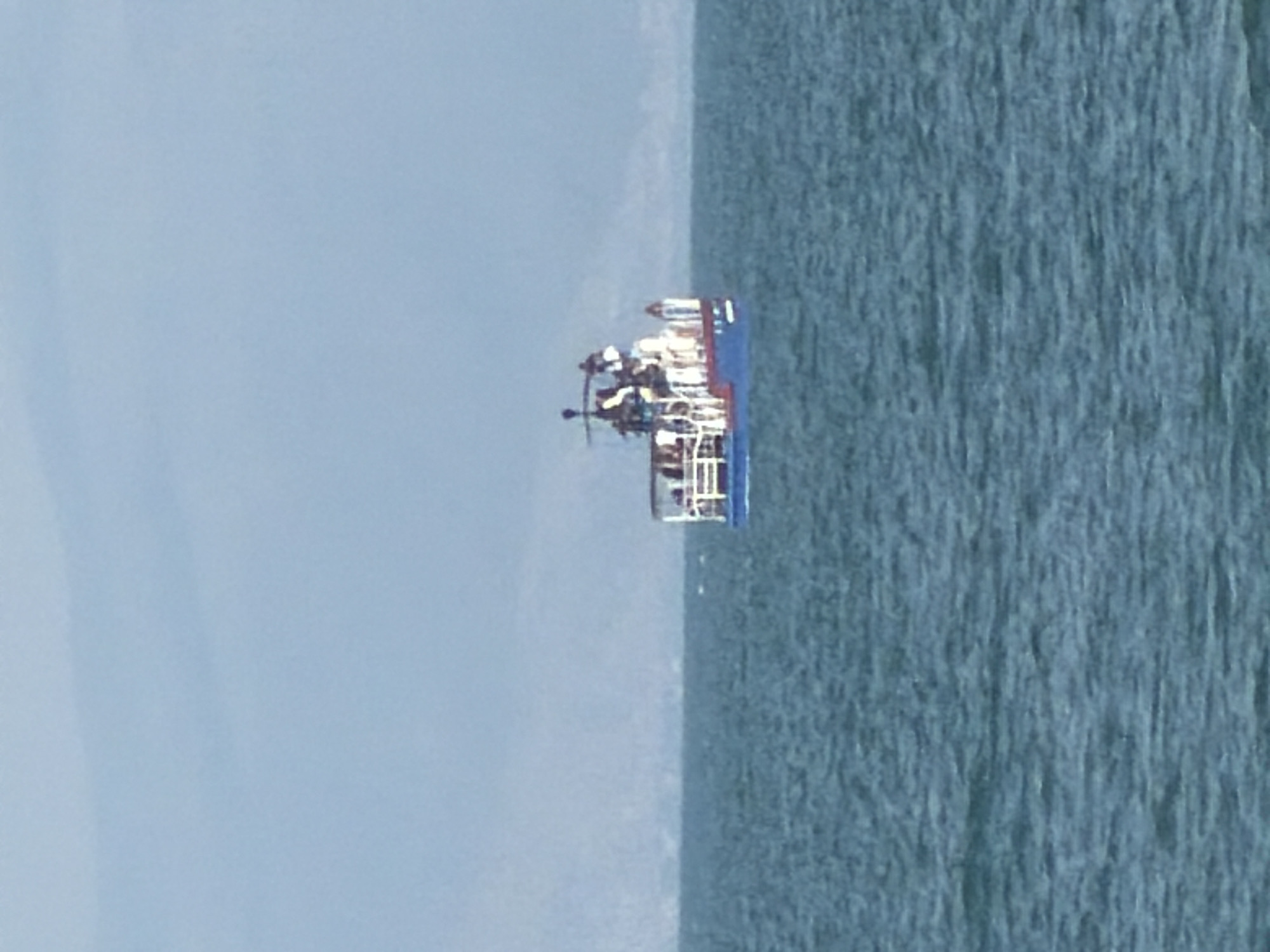 In the city of Tsamakaberd, Armenia, a small blue and white boat is visible on the water. The boat is carrying several passengers. The water is choppy, and the sky is overcast. In the distance, a faint outline of mountains can be seen. The overall impression is one of a calm day at sea.