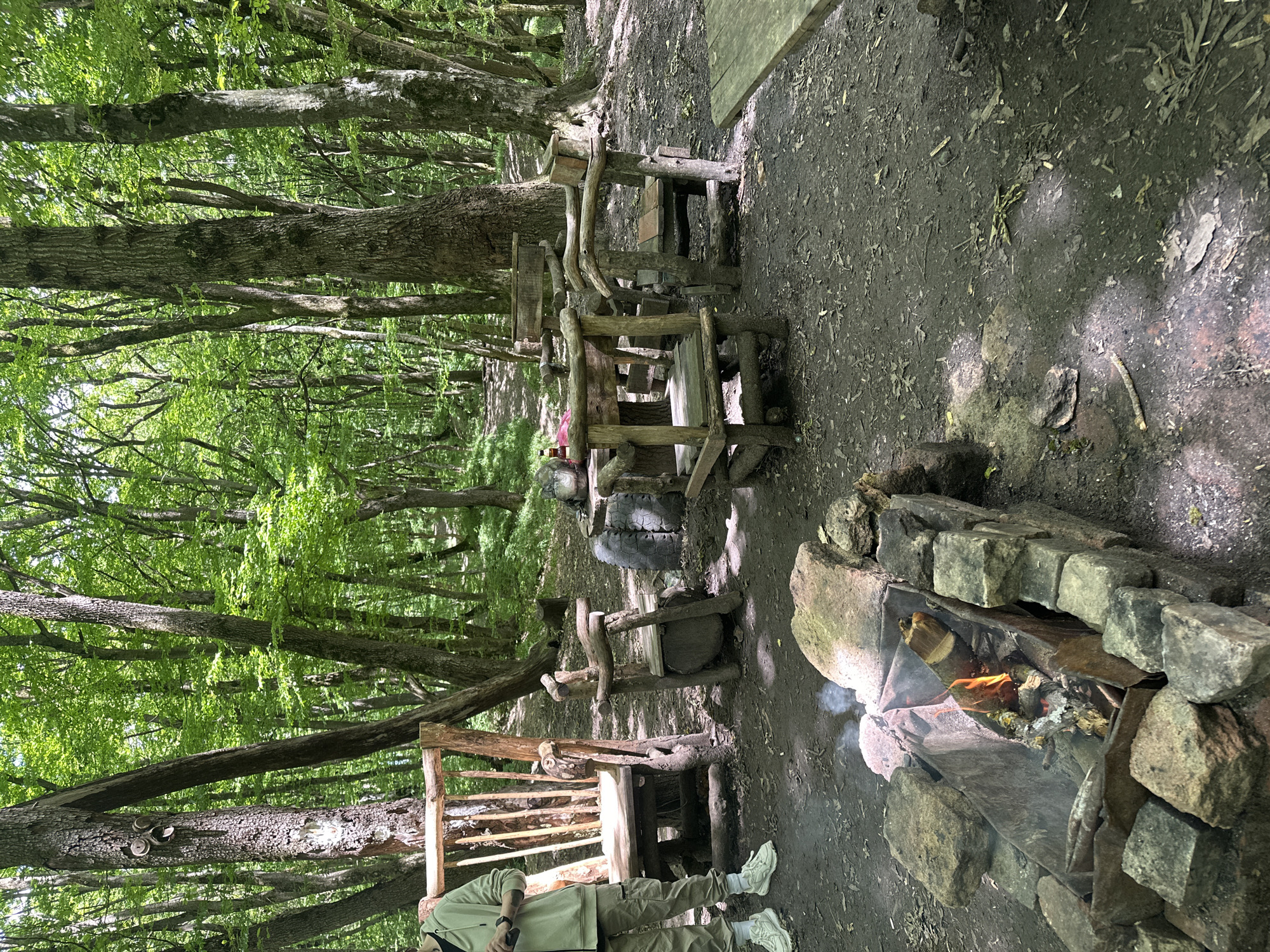 The image depicts a rustic outdoor campsite within a dense deciduous forest in Unknown, Unknown. A single individual is partially visible on the far left, standing and wearing a light-colored hooded sweatshirt, cargo pants, and white sneakers, holding a dark object.
The campsite features several handcrafted wooden furnishings. On the left, a large, high-backed wooden chair is positioned. To its right, a smaller wooden chair is visible. Further right, a grouping of three similar rustic wooden chairs surrounds a low, round table, partially constructed with what appear to be recycled tires at its base. Another flat wooden surface, possibly a table or bench, is partially visible on the far right.
In the foreground, a rectangular fire pit is constructed from stacked light-colored stones, enclosing a metal container. Wood is actively burning within the container, producing visible flames and light smoke.
The surrounding environment is a lush forest with numerous tall trees displaying vibrant green foliage, indicative of late spring or summer. The tree trunks are predominantly rough-barked and range in color from brown to gray. One tree trunk on the left has a carved or affixed face-like decoration. The ground consists of dark earth covered with scattered fallen leaves, twigs, and small debris. Dappled sunlight filters through the tree canopy, creating varied lighting across the scene. A dirt path is visible leading into the background forest.