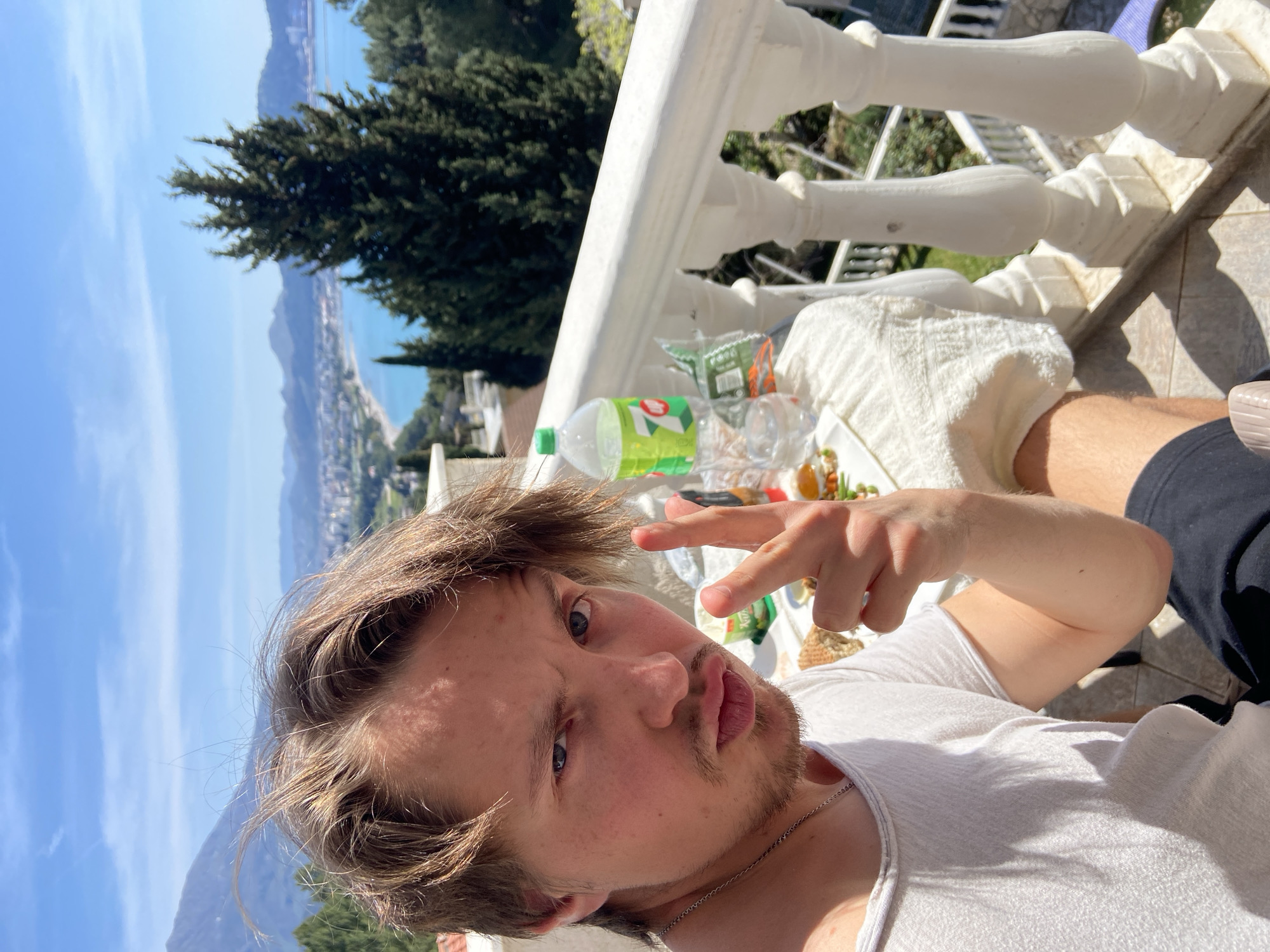 A young Caucasian man is taking a selfie on a balcony, likely in Šušanj, Montenegro, given the visible coastline and mountainous terrain in the background. He has light brown hair, a short beard, and is wearing a white t-shirt and a silver necklace. He is making a "peace sign" with his hand while puckering his lips. On the balcony, next to him, is a white railing and a spread of food and drinks including a bottle of 7-Up, a plastic cup, and some snacks on a white towel. A tree is visible behind the railing, and beyond that is a view of the sea and a city nestled along the coast. The sky is blue with some clouds. The image appears to have been taken on a sunny day.