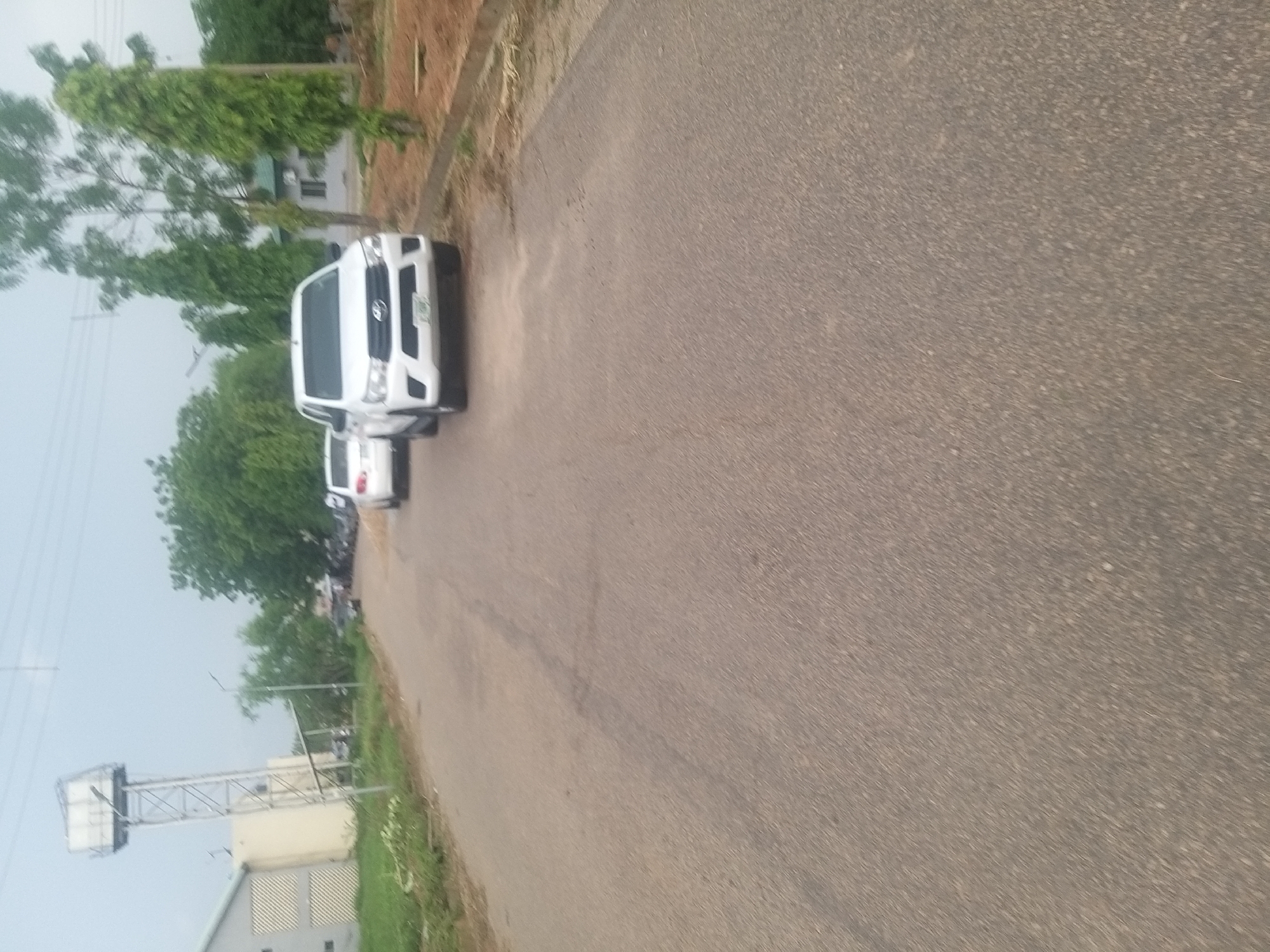The image depicts a view of a paved road in Kano, Nigeria, with two white vehicles present. The road surface is asphalt, appearing somewhat worn with visible seams. On the left side of the road, a large, industrial-style building is visible, adjacent to a tall, metal water tower. Green vegetation, including trees and grass, lines the sides of the road. In the background, more trees and buildings can be seen, suggesting an urban or semi-urban environment. The sky is overcast with a pale, diffused light, indicating it may be morning or late afternoon. The overall scene suggests a functional, possibly institutional, area within Kano.
