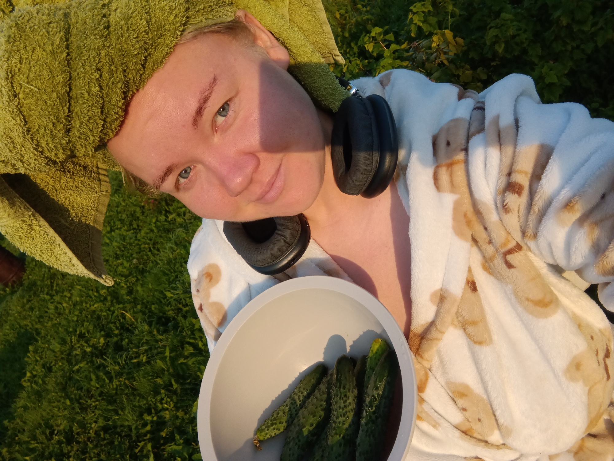 A close-up, high-angle selfie shows a person's face and upper body. The person has light skin, blue eyes, and light-colored eyebrows. They are wearing a green towel wrapped around their head and a white bathrobe with a light brown pattern of what appear to be cartoon bears. Black headphones are draped around the person's neck, with one earcup resting on their shoulder. In the foreground, the person holds a light gray bowl containing several cucumbers. The background consists of green grass and foliage, illuminated by warm, natural light, suggesting an outdoor setting, possibly in Foki, Russia, during the golden hour.