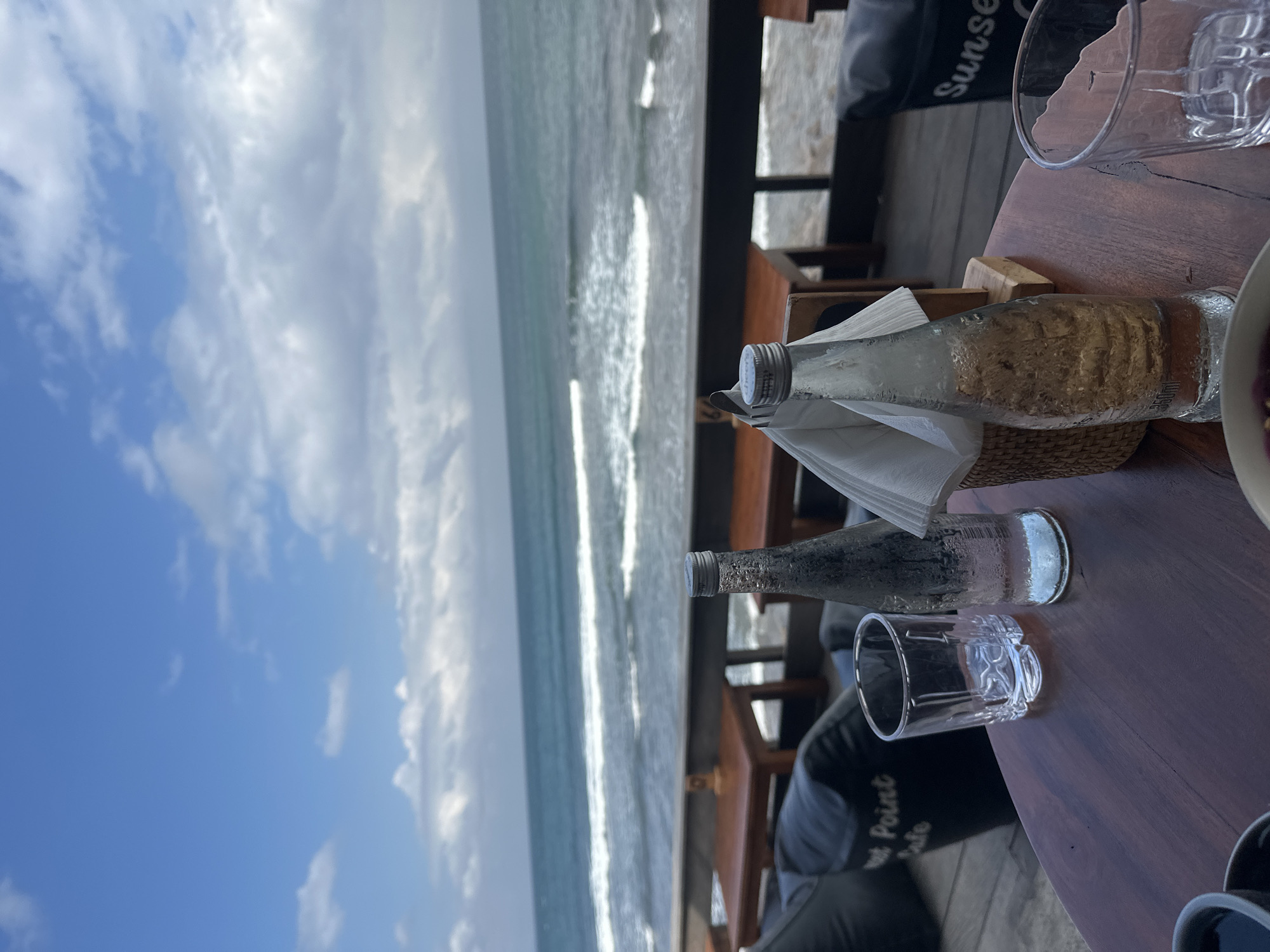 The image depicts a table at a beachfront cafe in North Kuta, Indonesia, overlooking the ocean. On the dark wooden table, there are two glass bottles of water, one mostly full and the other half-full, with condensation on the outside. A clear glass tumbler is positioned to the left of the fuller bottle, and another empty glass tumbler is to the right of the half-full bottle. A woven basket with napkins is placed between the two bottles. In the background, the ocean stretches to the horizon under a sky with scattered clouds. Wooden tables and chairs, as well as dark beanbag chairs, are visible on a wooden deck. One beanbag chair has the text "Sunset" on it, and another has "Point Cafe" visible. The scene suggests a relaxed dining or lounging atmosphere with a view of the sea.