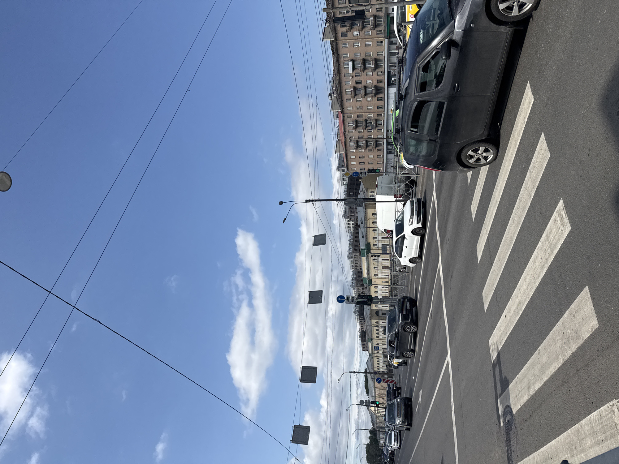 The image shows a street scene with a crosswalk in the foreground. Several cars are on the road, including a dark gray SUV on the right, a black SUV in the middle, and other vehicles further back. Buildings line the street in the background, and power lines and streetlights are visible overhead. The sky is blue with some scattered clouds. The overall scene suggests an urban environment on a clear day.